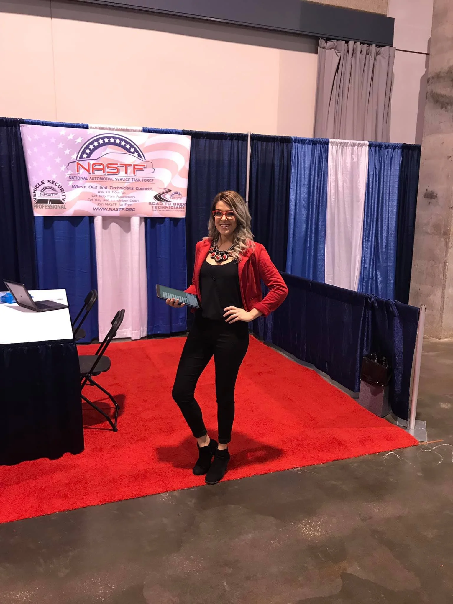 A woman with blond hair, wearing glasses, a black dress, and a red jacket, stands on a red carpet in front of a blue and white curtain backdrop. There is a NASTF banner behind her, indicating an automotive-related event.