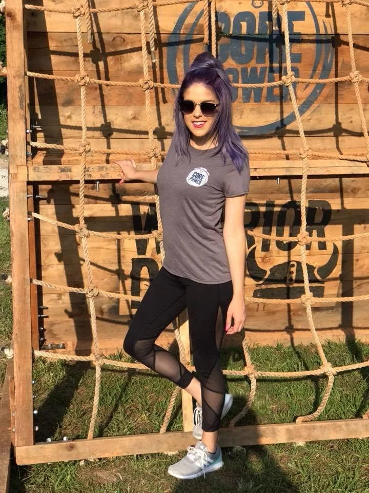 A woman with purple hair and sunglasses standing in front of a wooden and rope obstacle course with 'Core Power' and 'Warrior' signs in the background.
