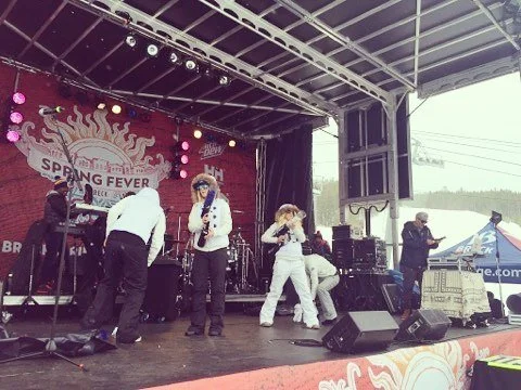 A group of people preparing for or performing on a stage at an outdoor event called 'Spring Fever' with musical instruments and sound equipment.