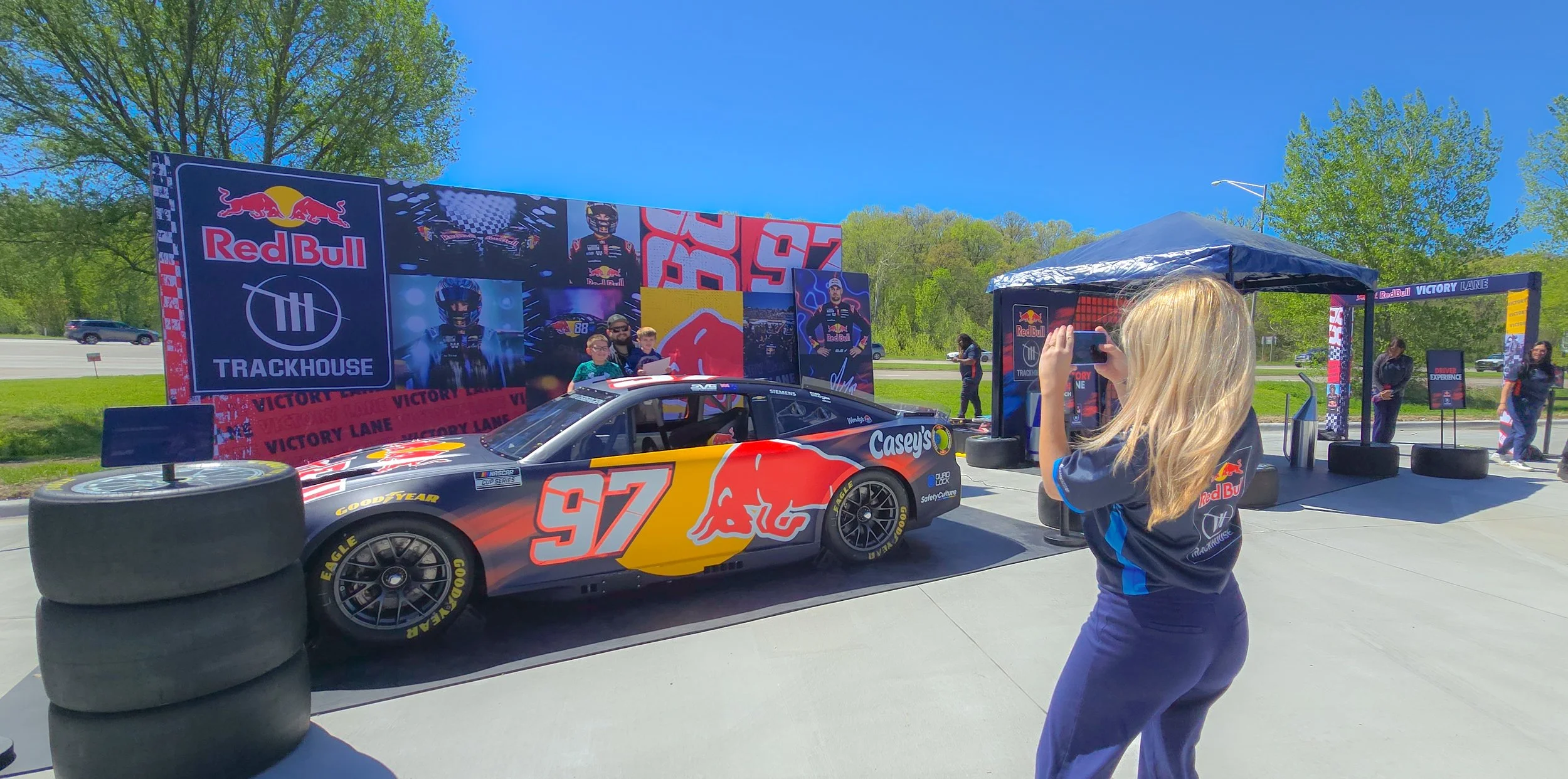 A racing car with the number 97 and Red Bull branding is parked on a track during a racing event. A woman with blonde hair is taking a photo of the car, and there is a large Red Bull banner with images of drivers and the event's name, Victory Lane, in the background. Trees and a highway are visible in the distance.
