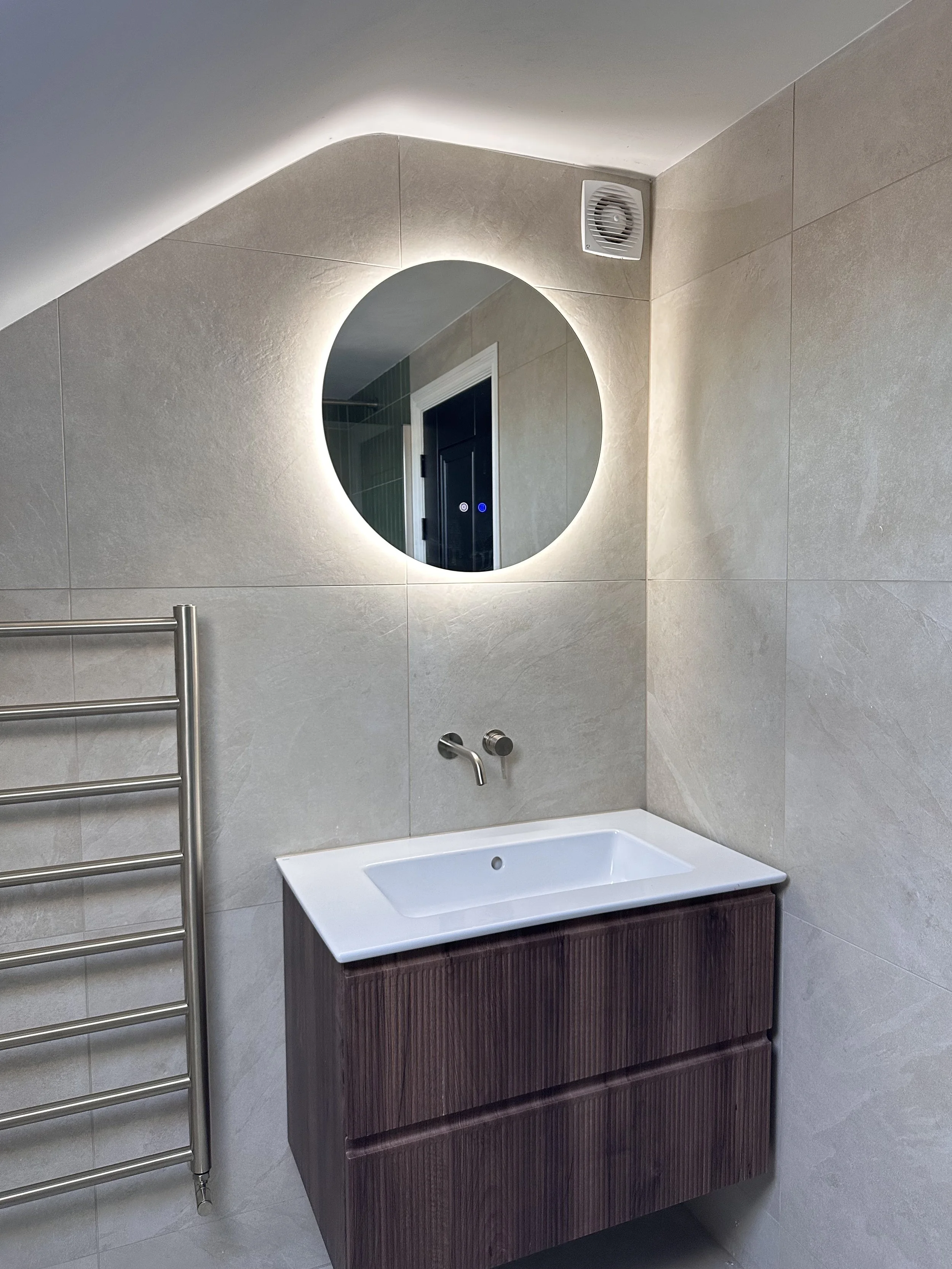 Modern bathroom with beige tile walls, a circular mirror with backlight, a white sink with a dark wood vanity, a wall-mounted faucet, and a stainless steel towel rack on the left.