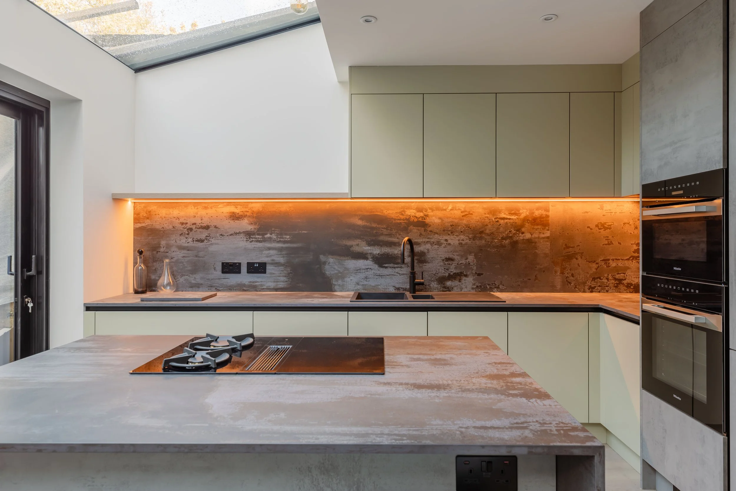 Modern kitchen with beige cabinets, black sink, gray and rust-colored backsplash, and built-in oven, with a skylight overhead.