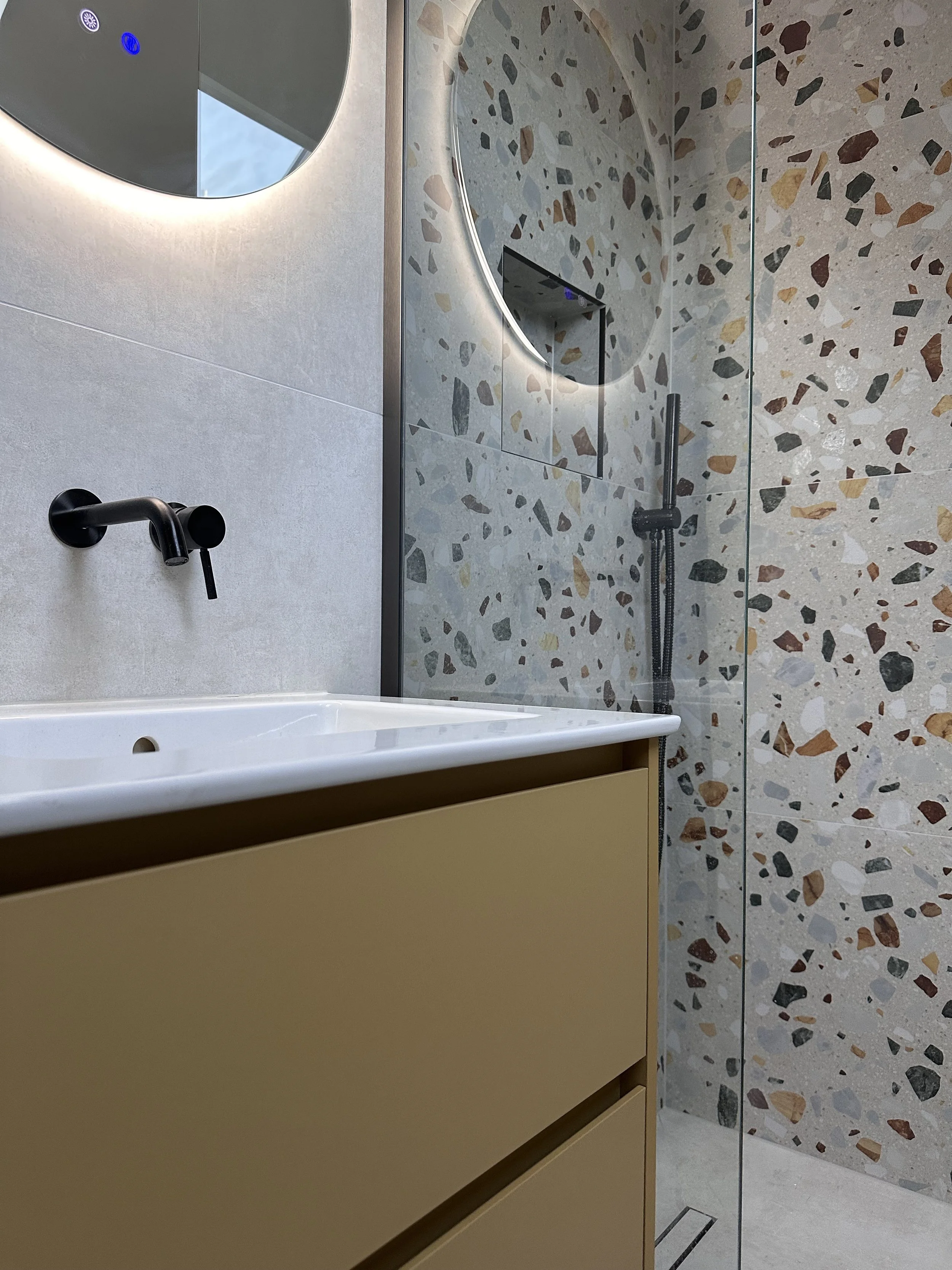 Modern bathroom with beige vanity, black faucet, round mirror with backlighting, terrazzo wall, and walk-in shower with black fixtures and glass partition.