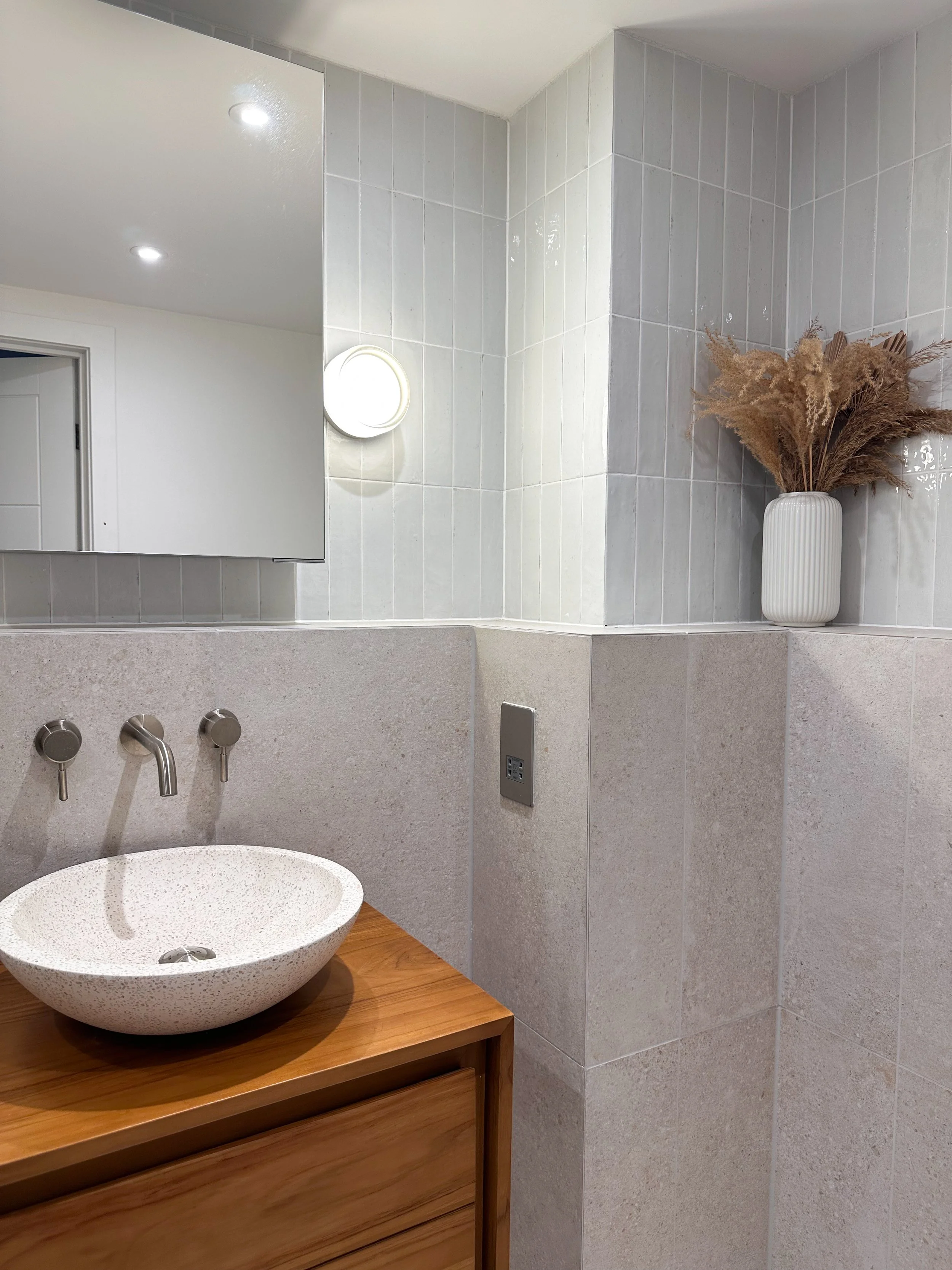 A modern bathroom with a large mirror, a white vessel sink on a wooden vanity, a wall-mounted faucet, a white vase with dried beige flowers, and tiled walls.