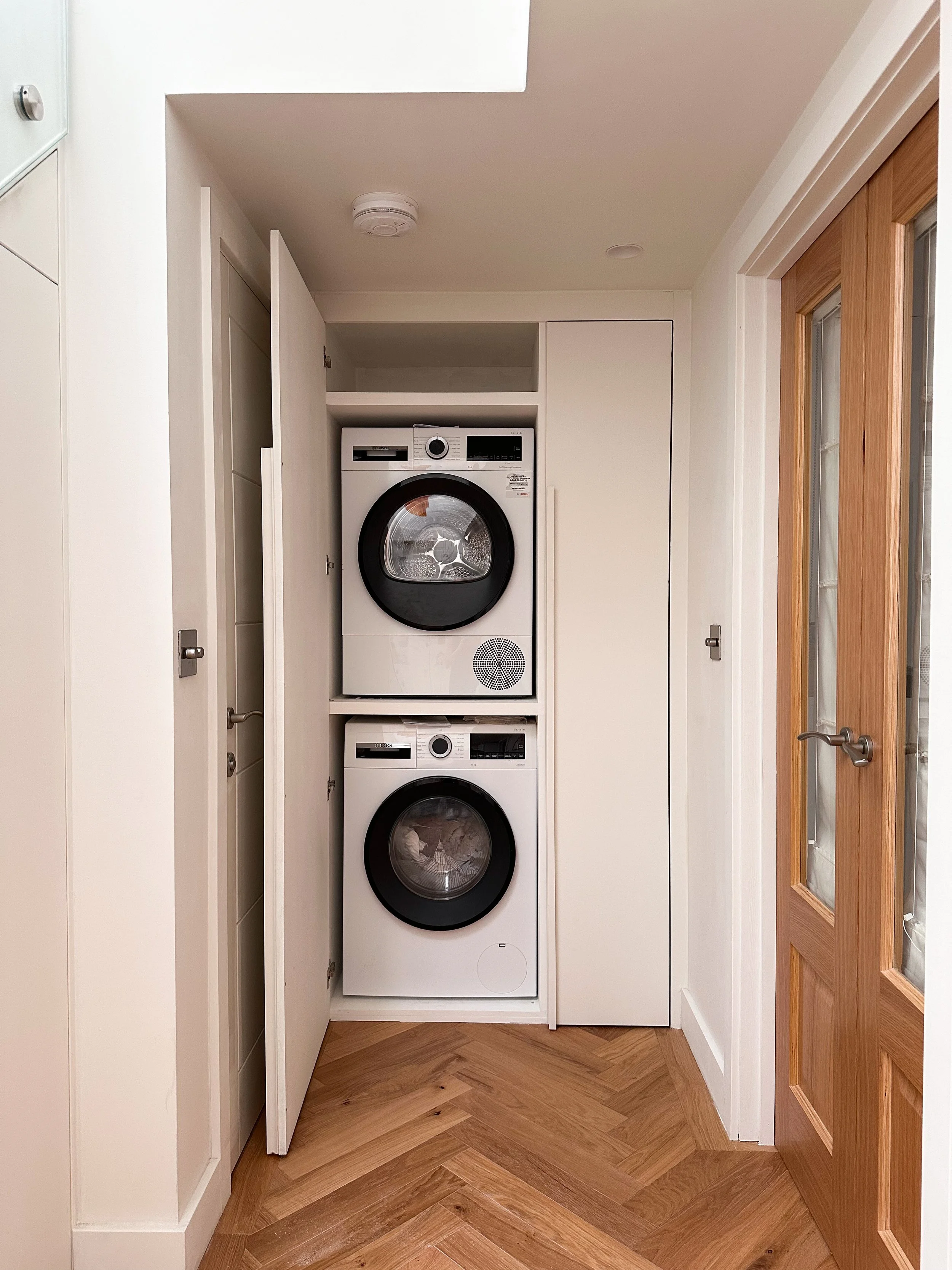 Stacked washer and dryer enclosed within cabinet doors in a laundry closet, with wooden door on the right and white walls.