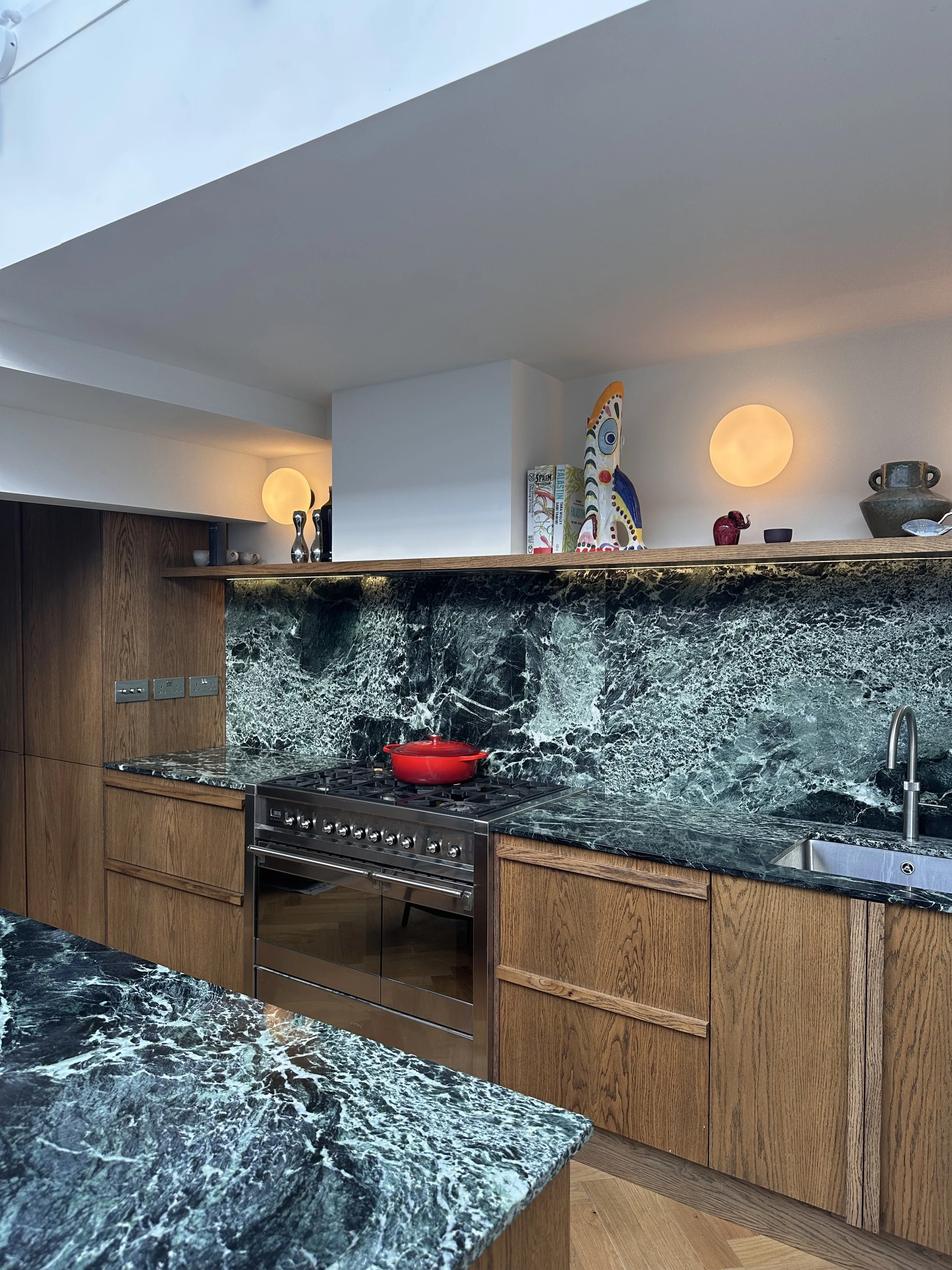 Modern kitchen with green marble countertops, wooden cabinets, a stainless steel stove with a red pot on top, and a white sink. Decorative items on the back shelf include art pieces, bowls, and sculptures, with two round wall lights illuminating the 