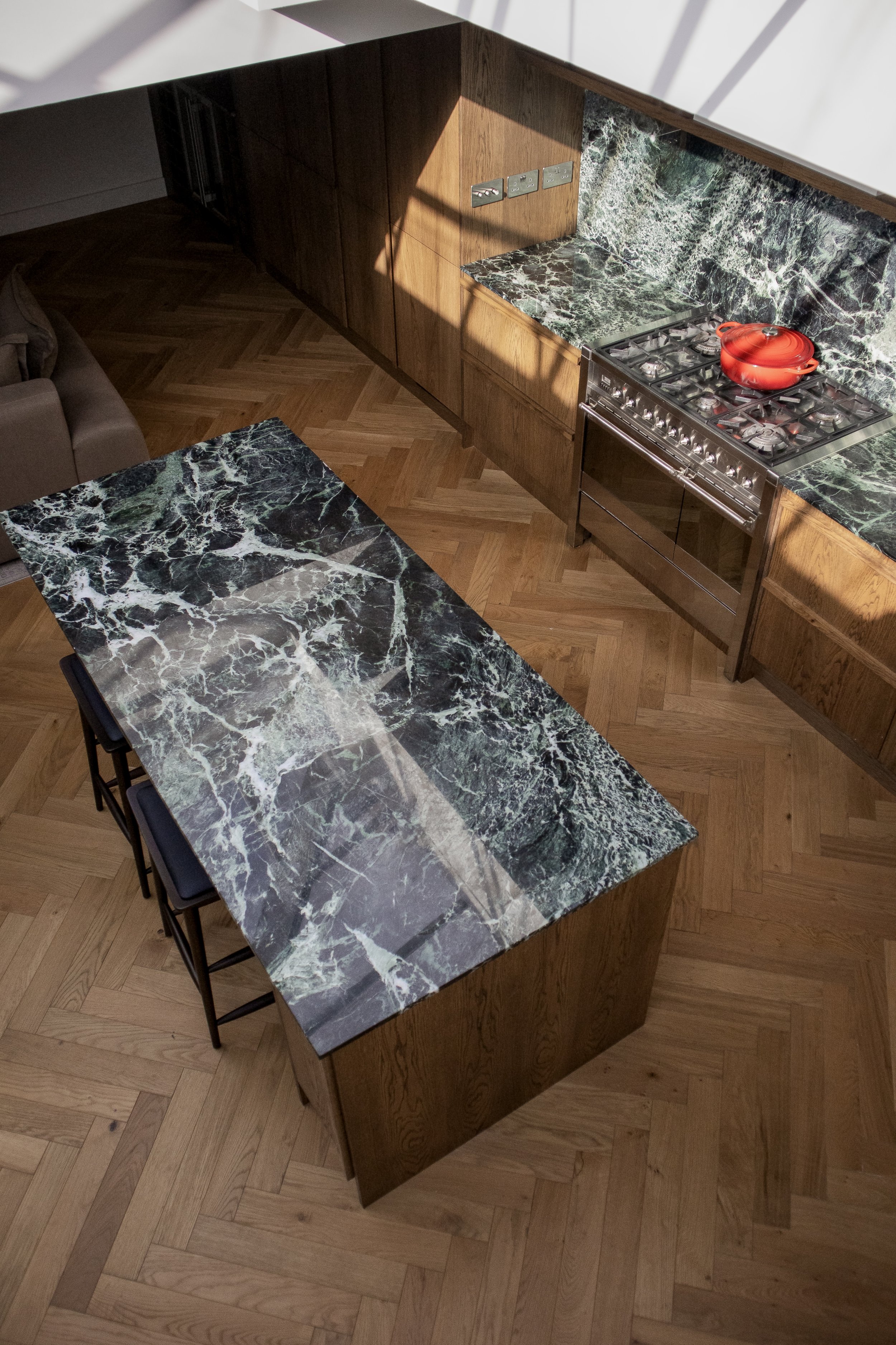 A top-down view of a modern kitchen with a wood floor, wooden cabinets, a marble countertop and backsplash, a stove with a red pot, and a marble-top kitchen island with three chairs.