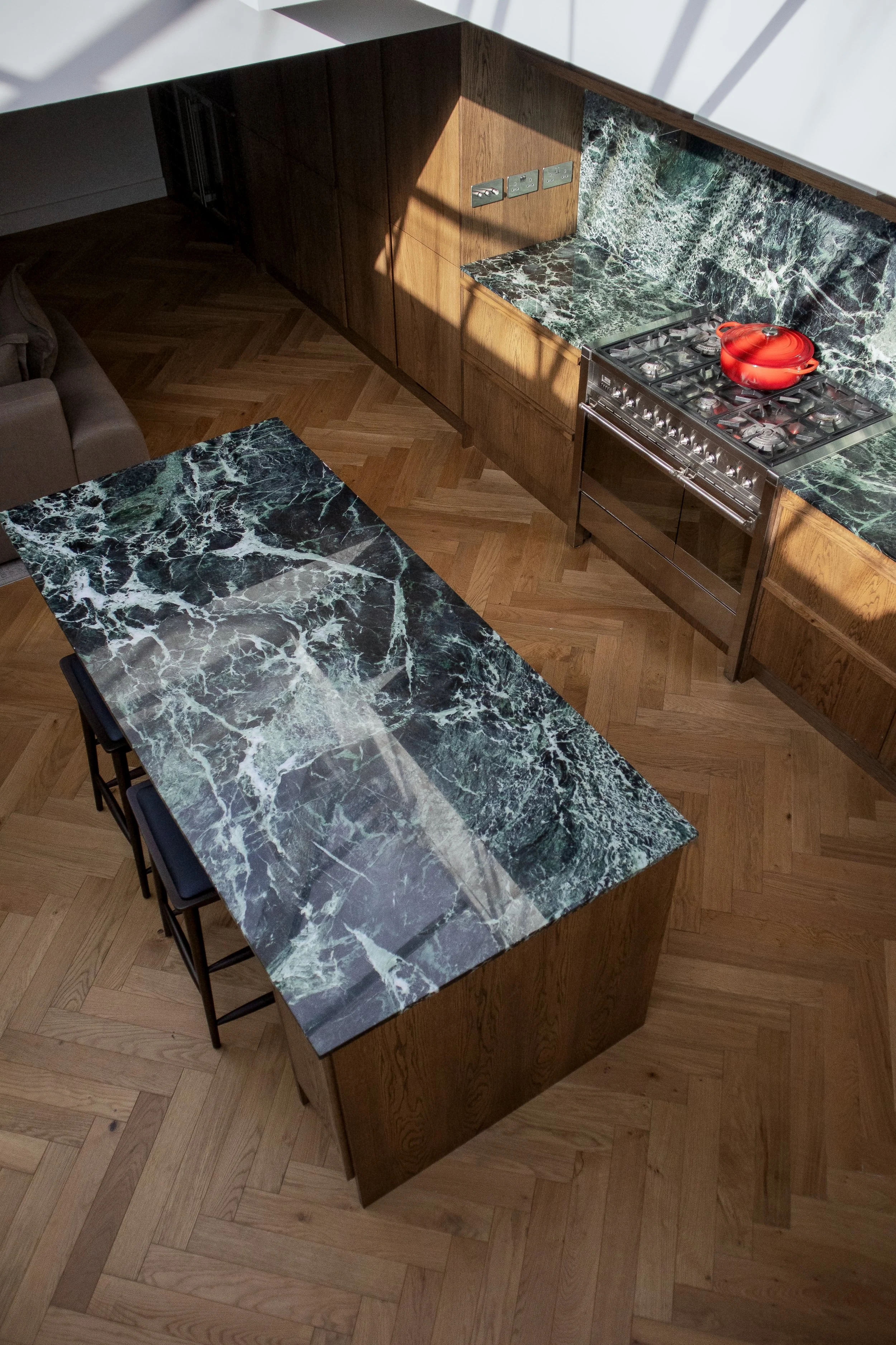 A kitchen with a black and green marble countertop on the island and walls, wooden cabinets, a stainless steel stove with a red pot, and a hardwood floor.