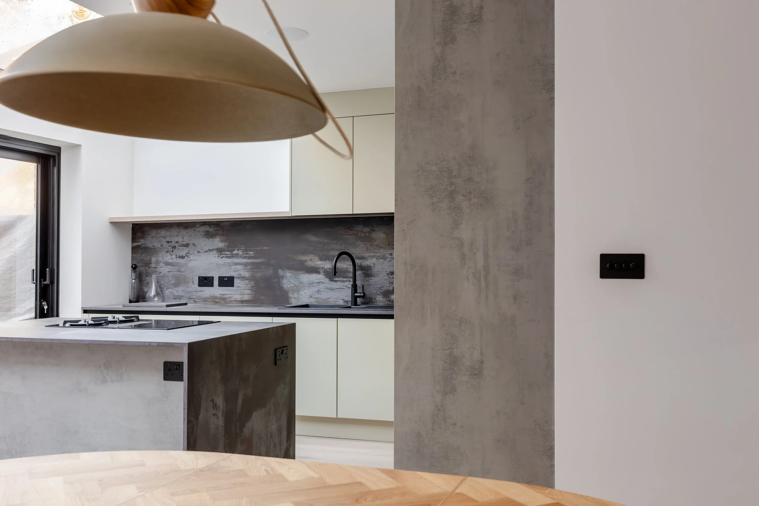 Modern kitchen with white cabinets, black faucet, textured gray backsplash, and a concrete-textured wall. An out-of-focus wooden table and a hanging lamp are also visible.
