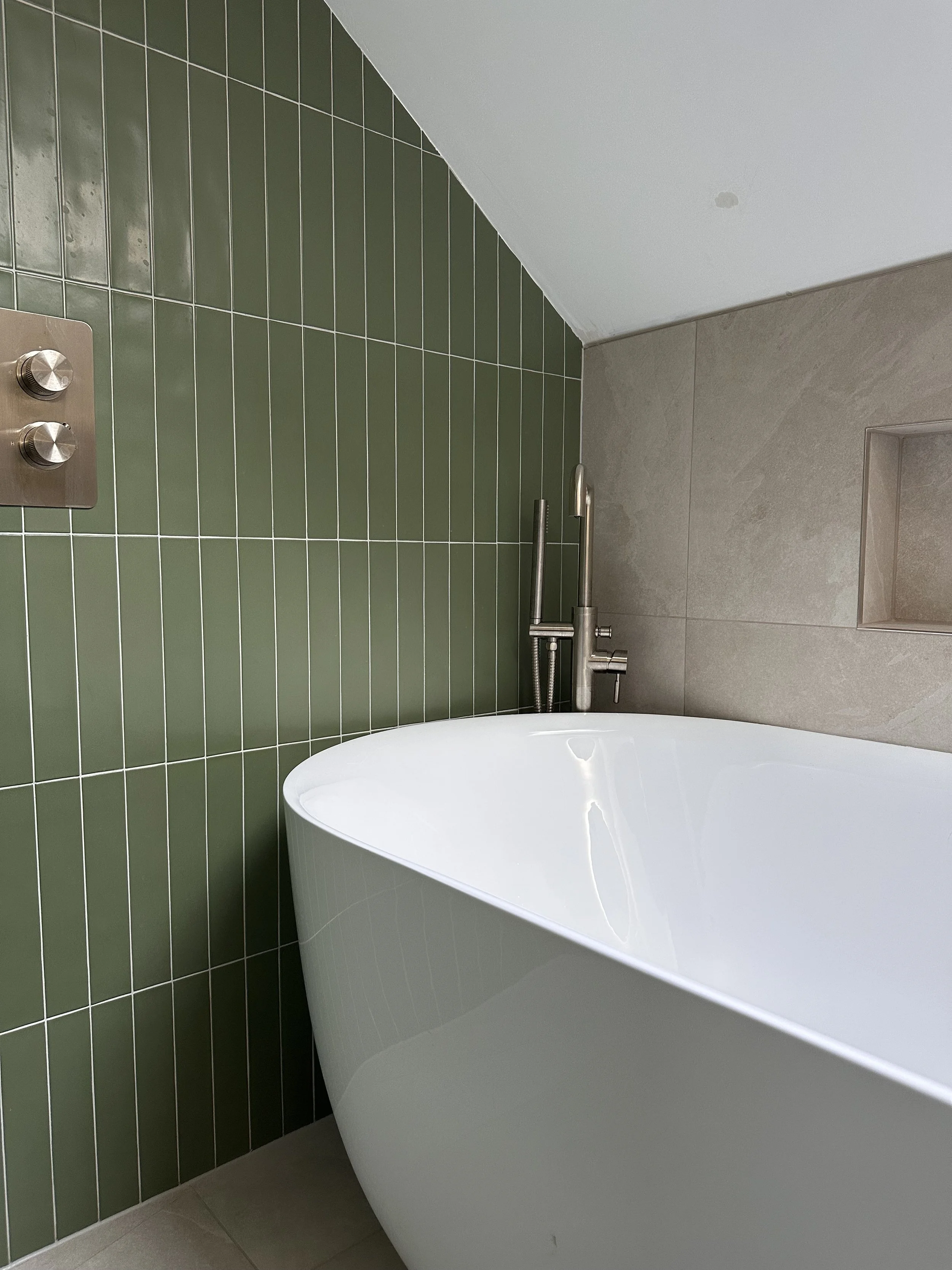 Modern bathroom with a white freestanding bathtub, green tiled wall, beige tiled wall with recessed shelf, and industrial-style faucet.