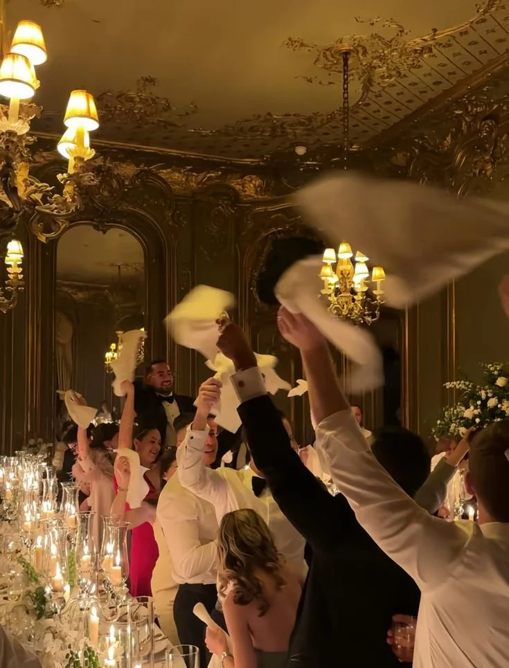 Guests celebrating at a wedding reception, waving napkins, in an opulent decorated room with chandeliers and mirror panels.
