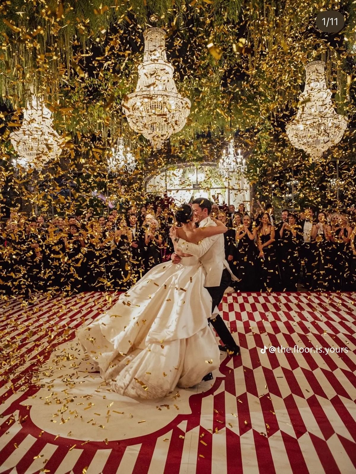 A bride and groom share a dance at their wedding reception, surrounded by guests, with gold confetti falling and chandeliers hanging from the ceiling.