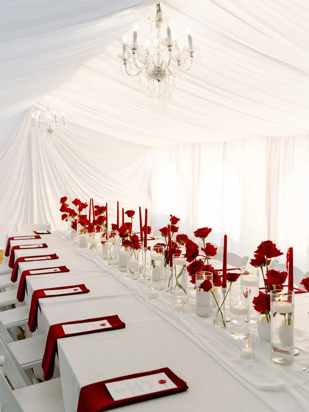 Elegant wedding reception table with white tablecloth, red napkins, and red floral arrangements in glass vases, with candles, set inside a white tent with draped fabric and chandeliers.