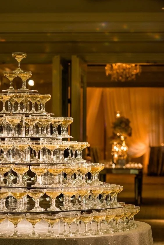 A pyramid of stacked champagne glasses arranged on a table in an elegant, warmly lit venue with draped curtains and floral arrangements in the background.