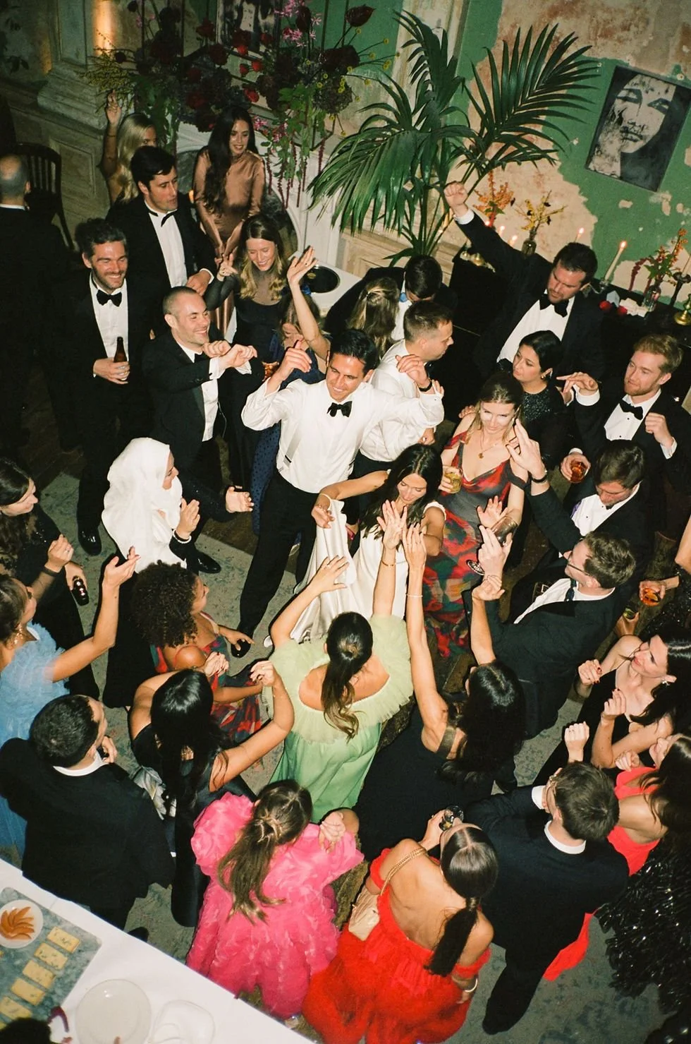 People dancing and celebrating at an indoor party, dressed in formal attire, with some holding drinks, in a decorated room with plants, candles, and artwork on the walls.