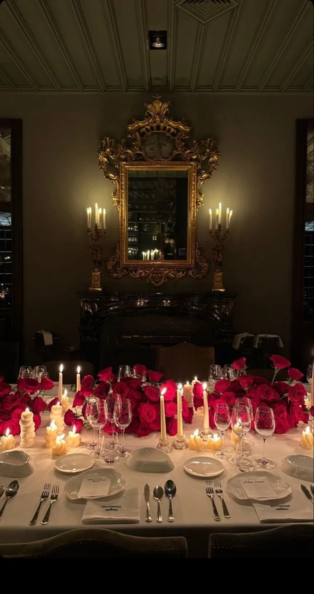 A elegantly decorated dining table set for a formal dinner with white plates, silverware, wine glasses, and white candles. The table features a large centerpiece of red roses and candles. Behind the table, a dark fireplace mantle and a large ornate gold-framed mirror are visible, with wall-mounted candle sconces on either side.