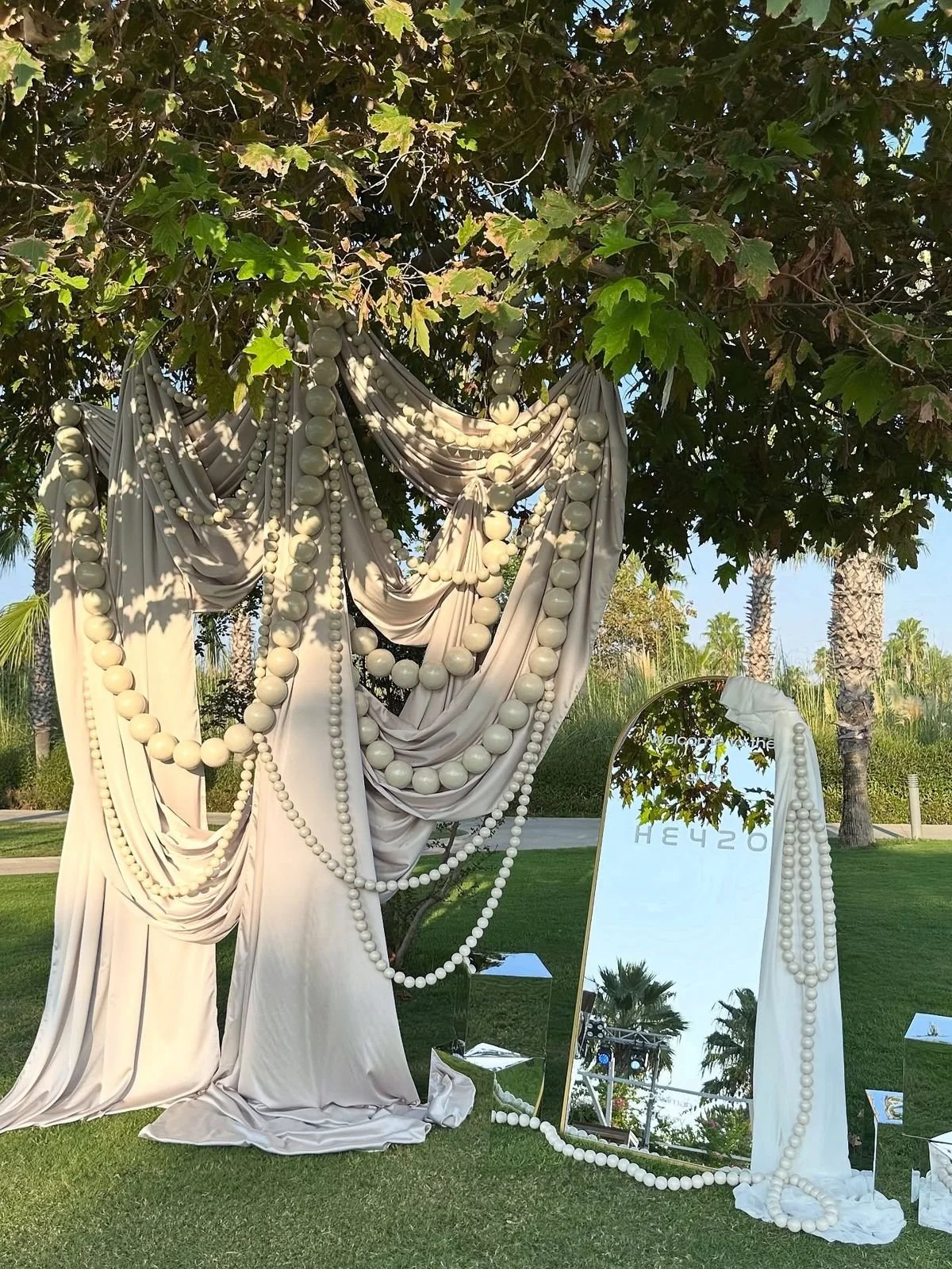 Decorative display with draped white fabric and multiple strands of large white beads hanging from a tree, with a mirror and a small sign, set in a landscaped outdoor area with palm trees and green grass.