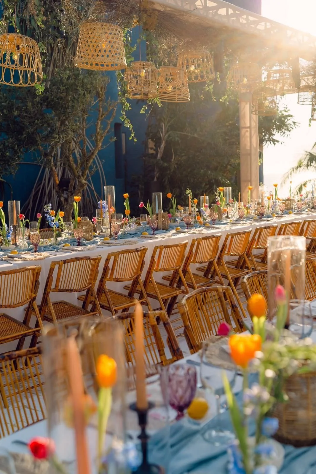 Long outdoor dining table decorated with colorful flowers, candles, and glassware, set for an event in a garden with hanging wicker lanterns and trees, bathed in warm sunlight.