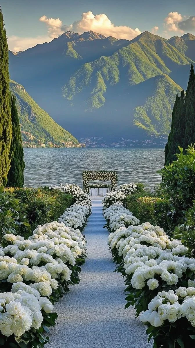 A wedding aisle decorated with white roses leading to a small wooden arch over a lake, with green mountains and a partly cloudy sky in the background.