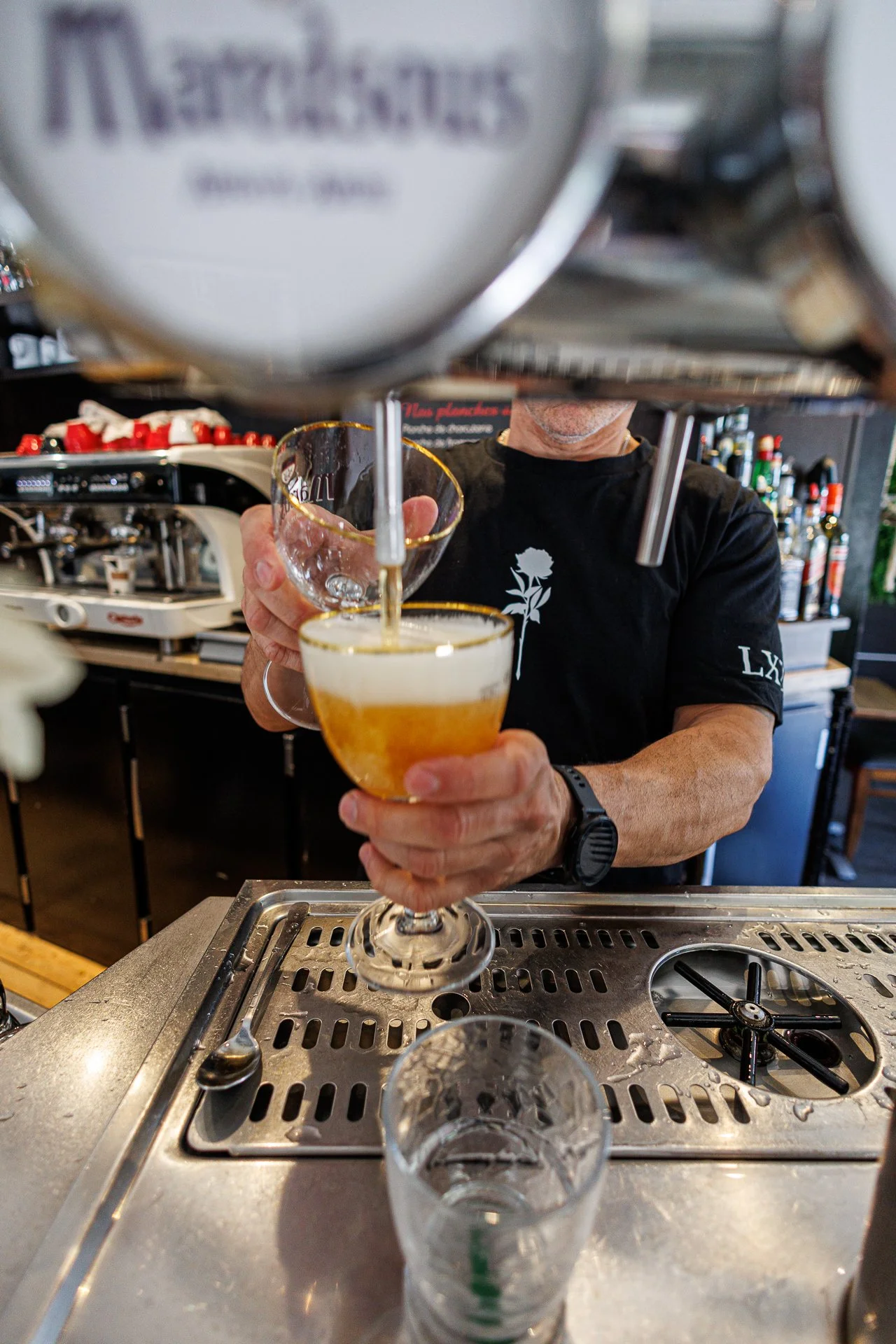 Un homme verse une boisson dans un verre à cocktail. Il porte une montre noire et un t-shirt noir avec une fleur blanche.