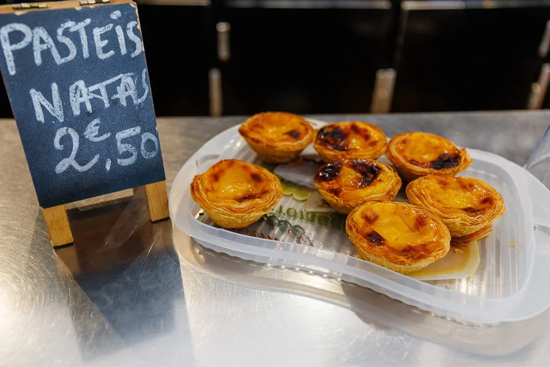 Plate de petites tartelettes à la crème ou à la confiture, sur un plateau en plastique. Un petit panneau à côté indique: 'Pastéis Nata 2,50 €'.