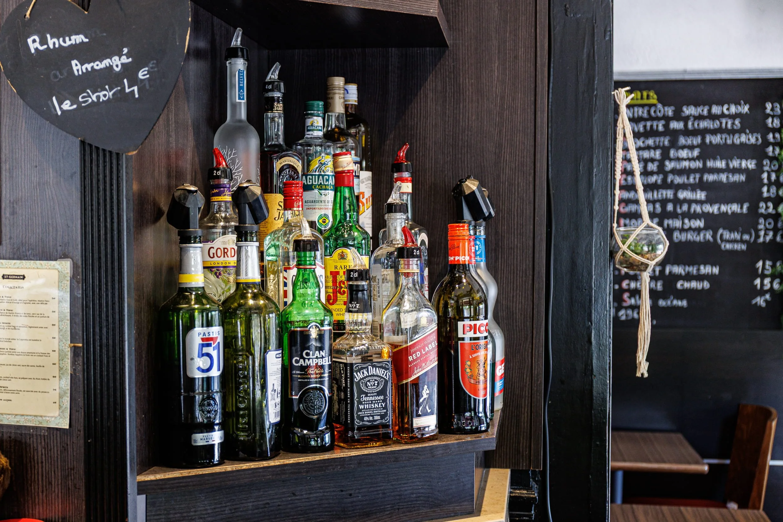Une étagère noire en bois avec plusieurs bouteilles d'alcool dont des spiritueux comme du whisky, de la vodka et du rhum, et un tableau noir avec une liste de plats en français.