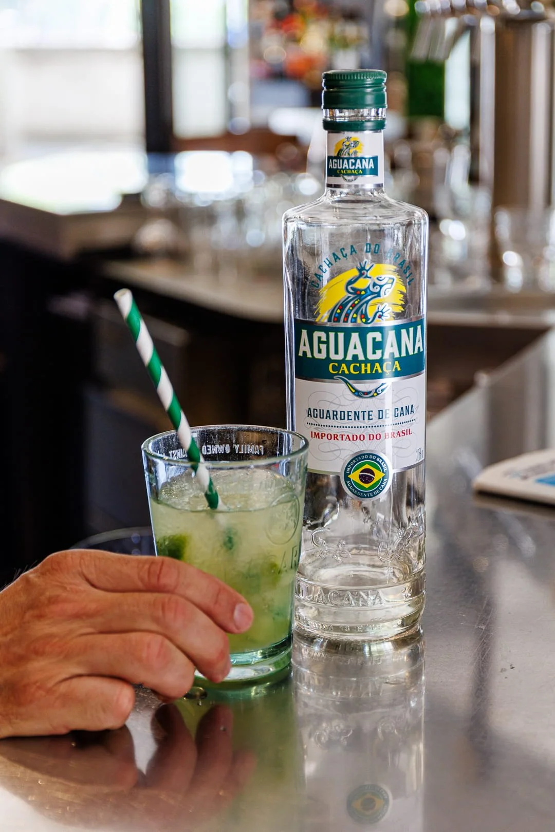 Une bouteille d'Aguacana Cachaça et un verre contenant une boisson verte avec des glaçons, une personne tient une paille rayée vert et blanc. Arrière-plan flou d'un bar ou cuisine.