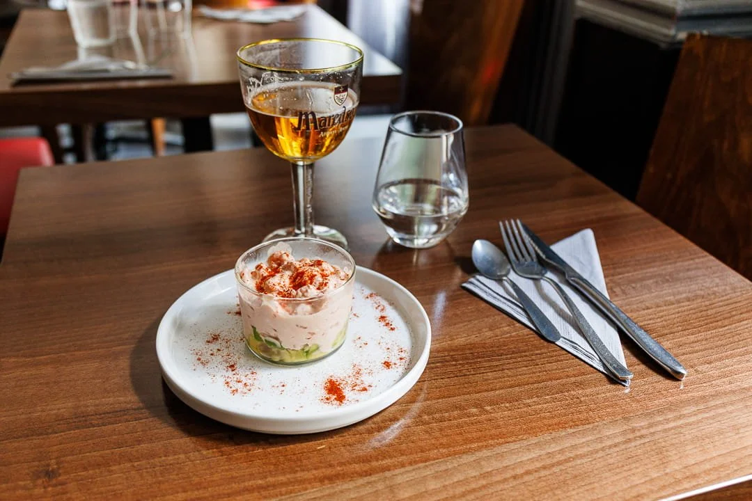 Verre de vin blanc, verre d'eau, petit plat avec un tartare de saumon, fourchette, couteau, cuillère, nappe en papier sur une table en bois dans un restaurant.