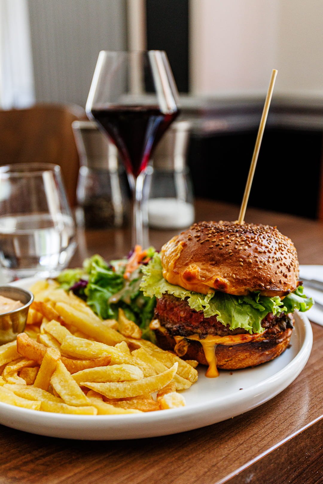 Un cheeseburger avec des frites, une salade, un verre de vin rouge, et un verre d'eau sur une table en bois.