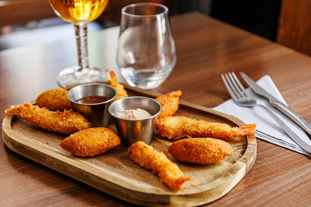 Plat de beignets de crevettes accompagnés de deux sauces, servie sur une planche en bois, avec un verre d'eau, un verre de vin, des couverts et une serviette en papier sur une table en bois.