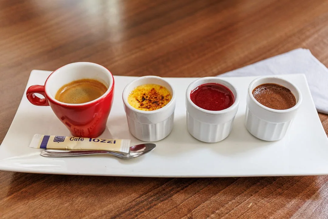 Assortiment de quatre petits contenants de sauces ou de desserts sur une assiette blanche, accompagnés d'une petite cuillère et d'un sachet de sucre, le tout placé sur une table en bois.