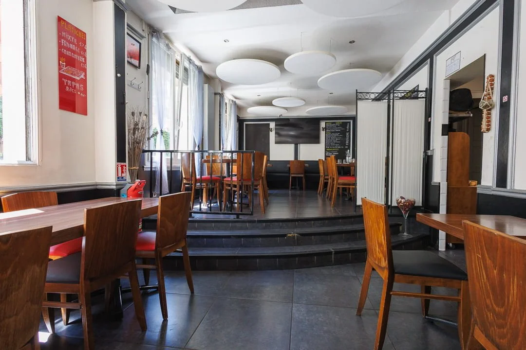Salle de restaurant avec tables en bois, chaises en bois, décor moderne, avec un petit escalier menant à une zone surélevée, à l'arrière un mur noir avec un tableau noir et un écran.