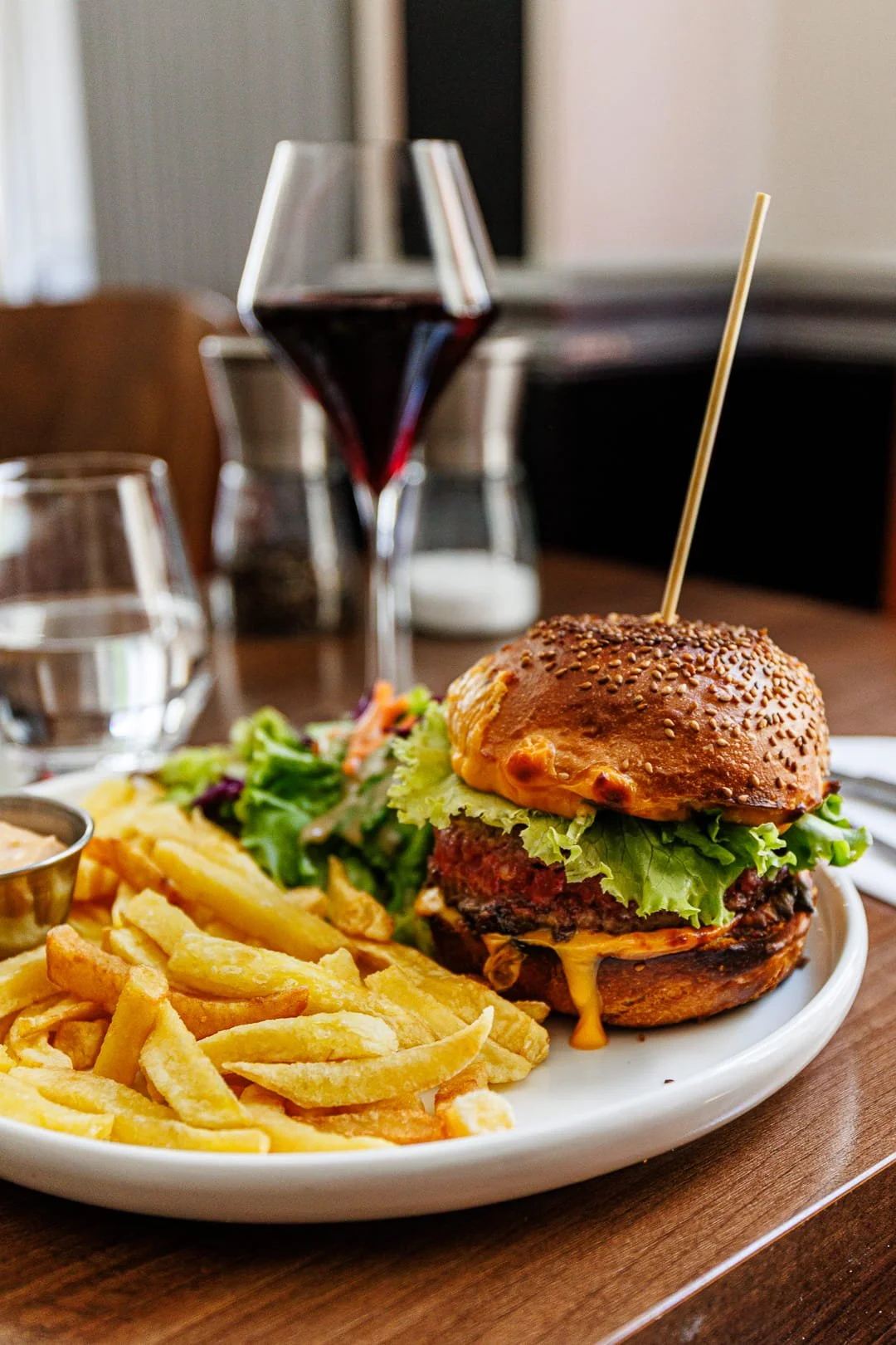 Un burger avec laitue, tomate, fromage, un steak, servi avec des frites, une salade et une sauce, accompagné d'un verre de vin rouge et une carafe d'eau sur une table en bois.