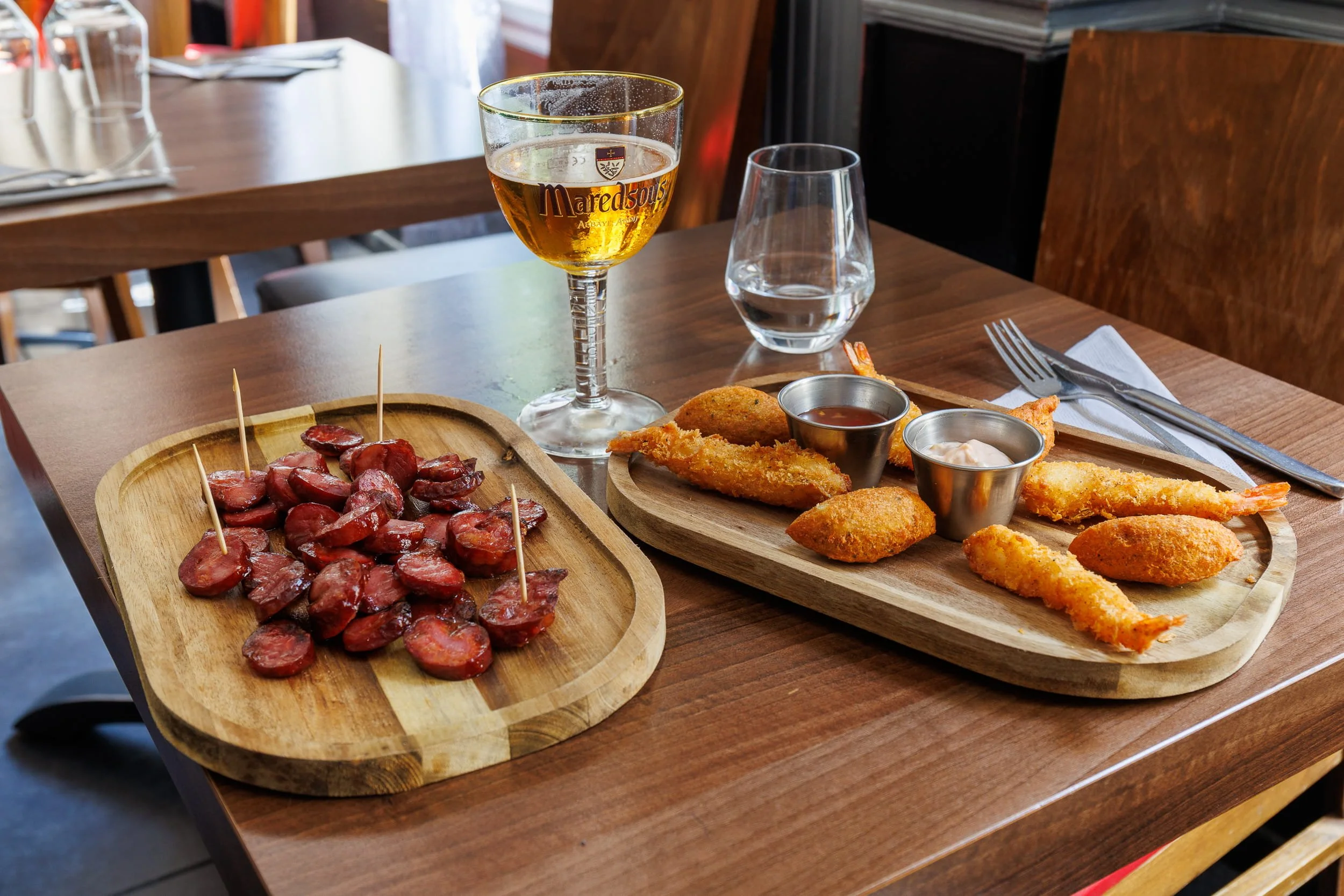 Une table avec un plateau en bois contenant des tranches de saucisses et des petites brochettes, à côté un autre plateau avec des filets de poisson panés et des sauces, avec un verre d'eau, une coupe de vin blanc, et un verre d'eau devant. En arrière