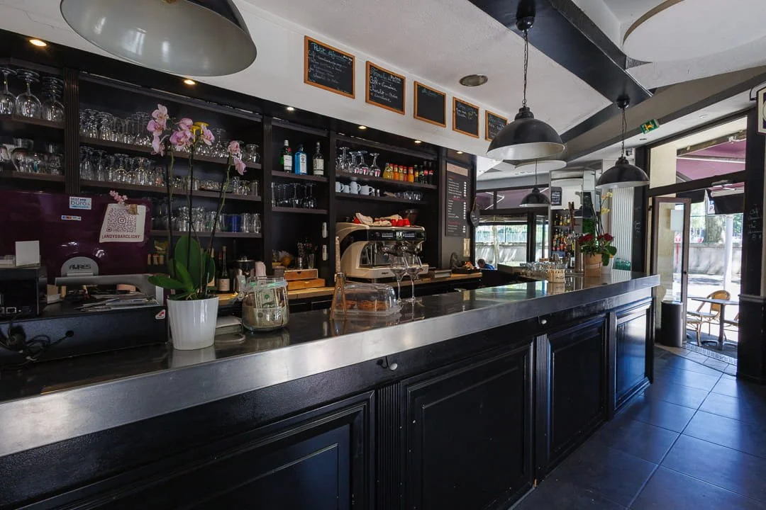 Bar de café avec étagères remplies de verres et bouteilles, comptoir en bois noir avec des plantes et des équipements de café, éclairage suspendu, devant une grande fenêtre donnant sur l'extérieur.