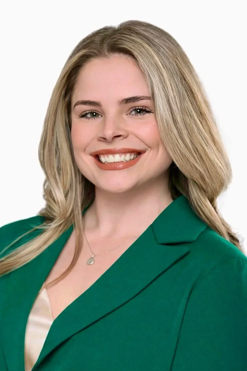 Headshot of a smiling woman with blonde hair, wearing a green blazer and a silver necklace, against a plain white background.