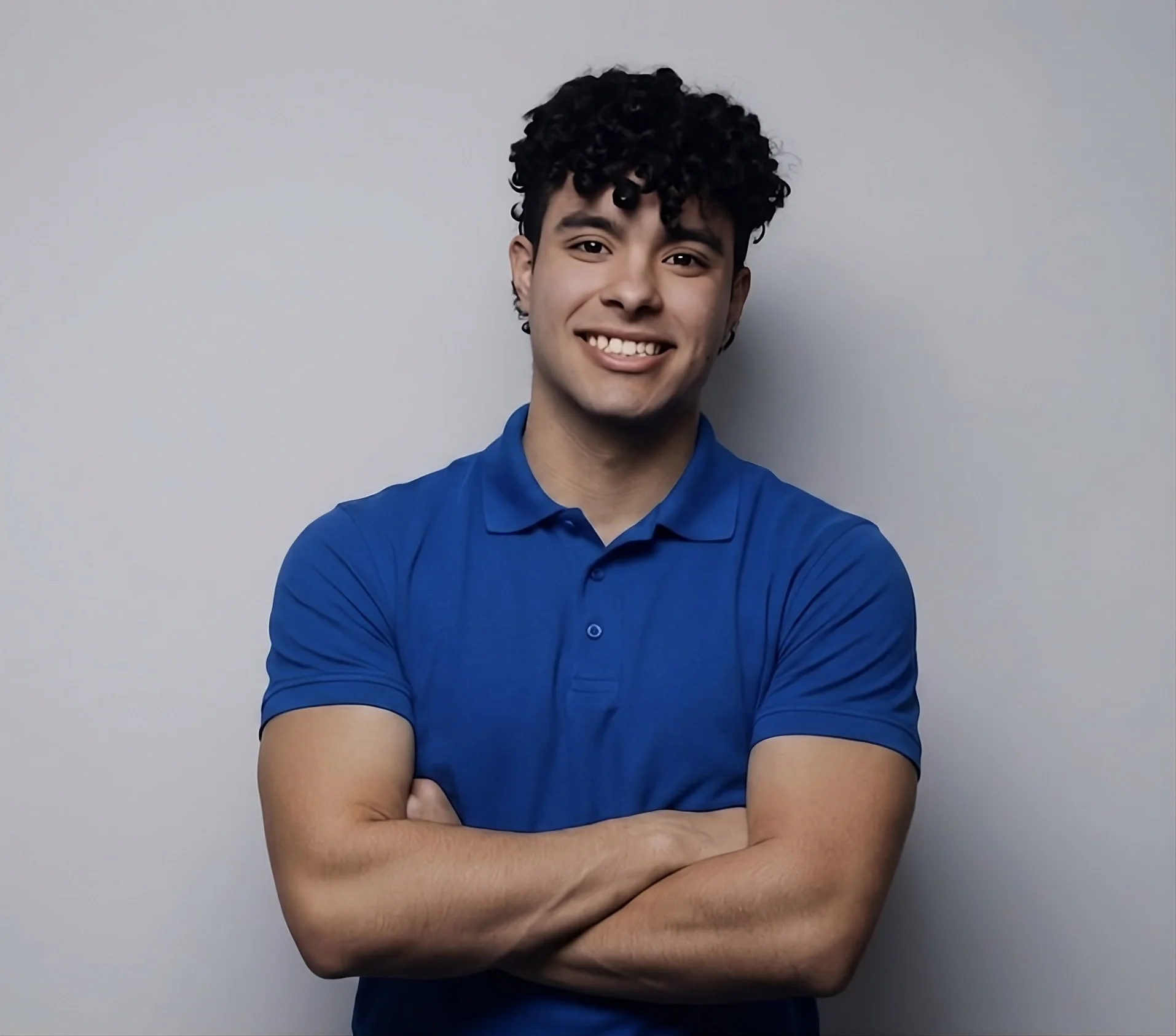 A young man with curly black hair, wearing a blue polo shirt, standing with his arms crossed and smiling against a plain gray background.