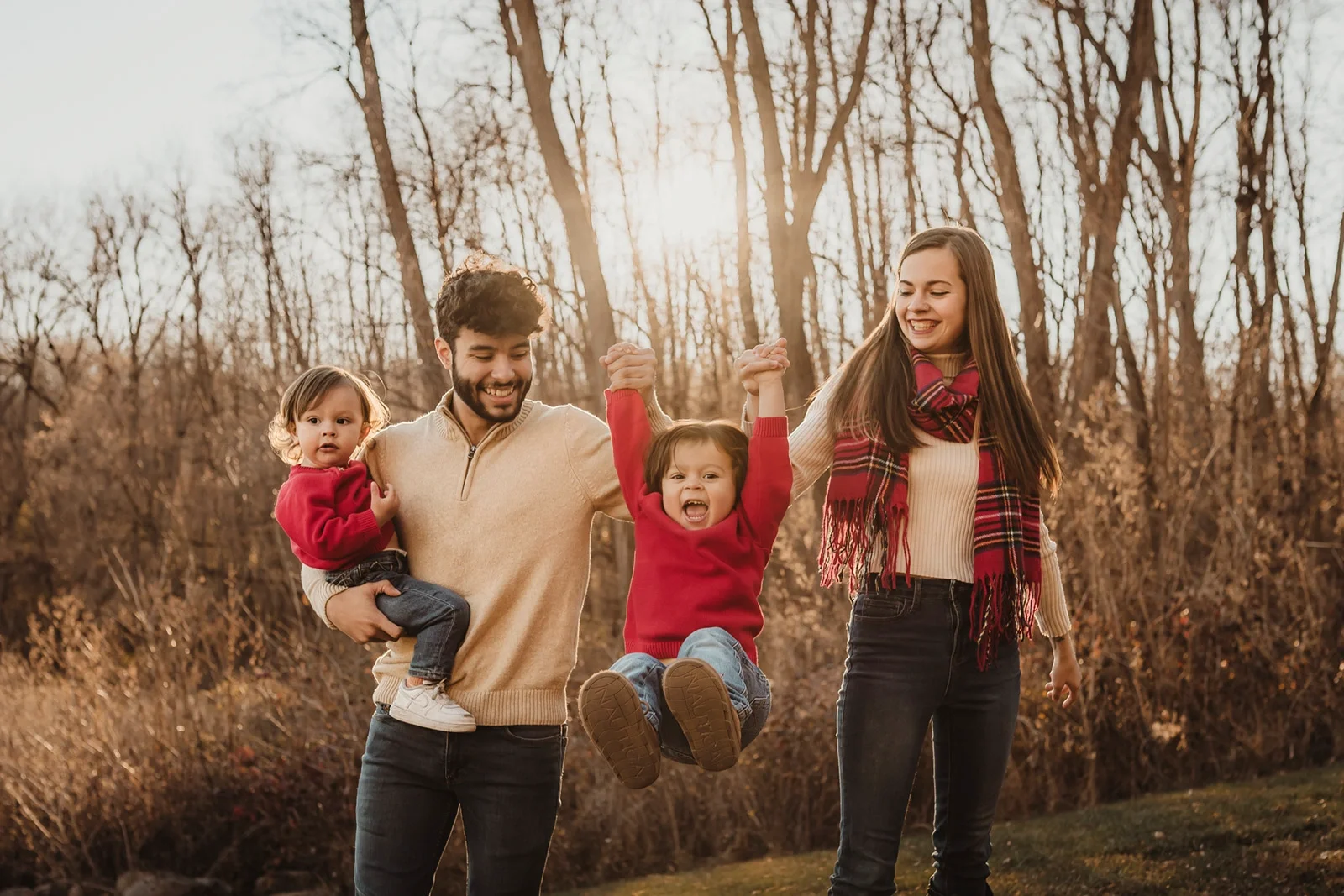 Family of four outdoors in a wooded area during fall, with two children, a father, and a mother, enjoying the moment. The father is holding a young girl, the mother is walking with a teenage girl, and a young boy is being lifted by the father, all smiling and happy.