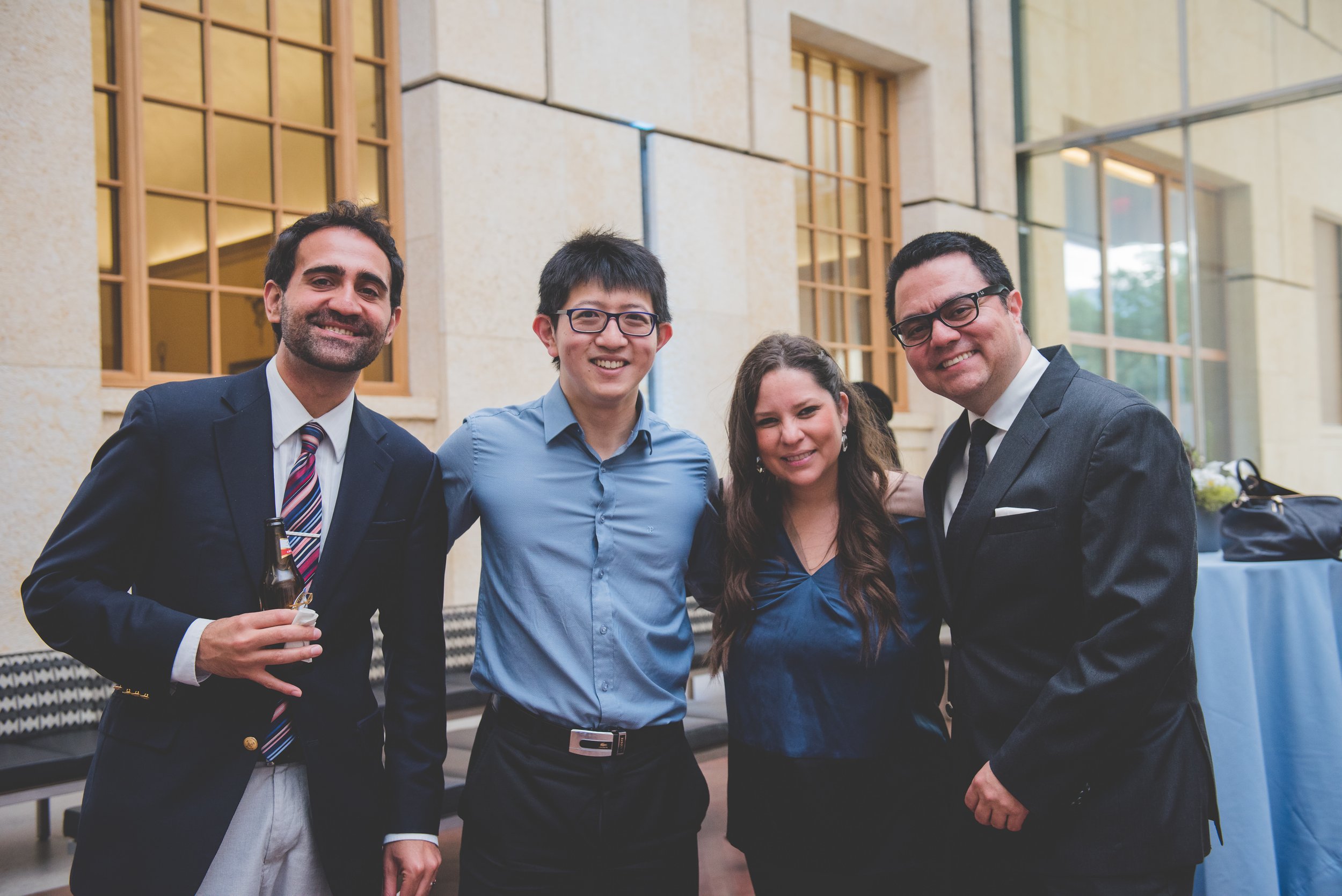 Four people dressed in formal attire, standing together outside a building with large windows and beige walls, smiling at the camera.