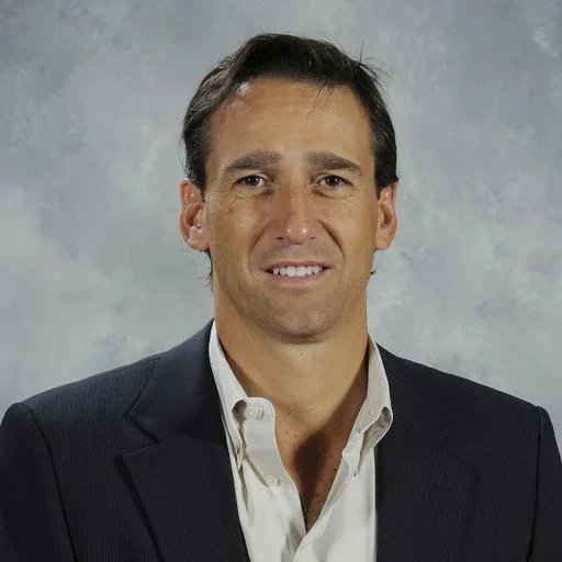 A man with dark hair and a slight smile wearing a dark suit jacket and white shirt against a plain background.