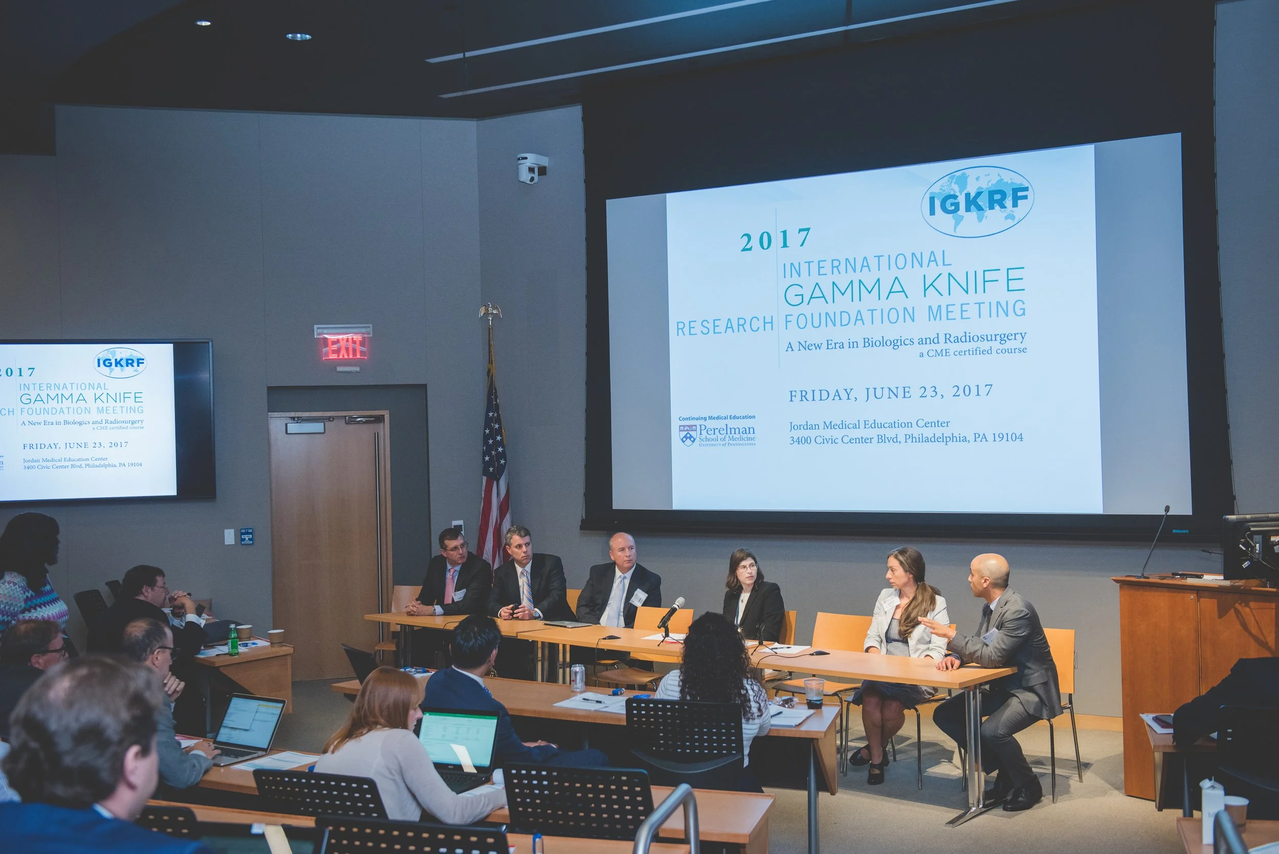 Conference room with panel of speakers at a research foundation meeting, large projection screen displaying event details, audience members seated at tables, and American flag.