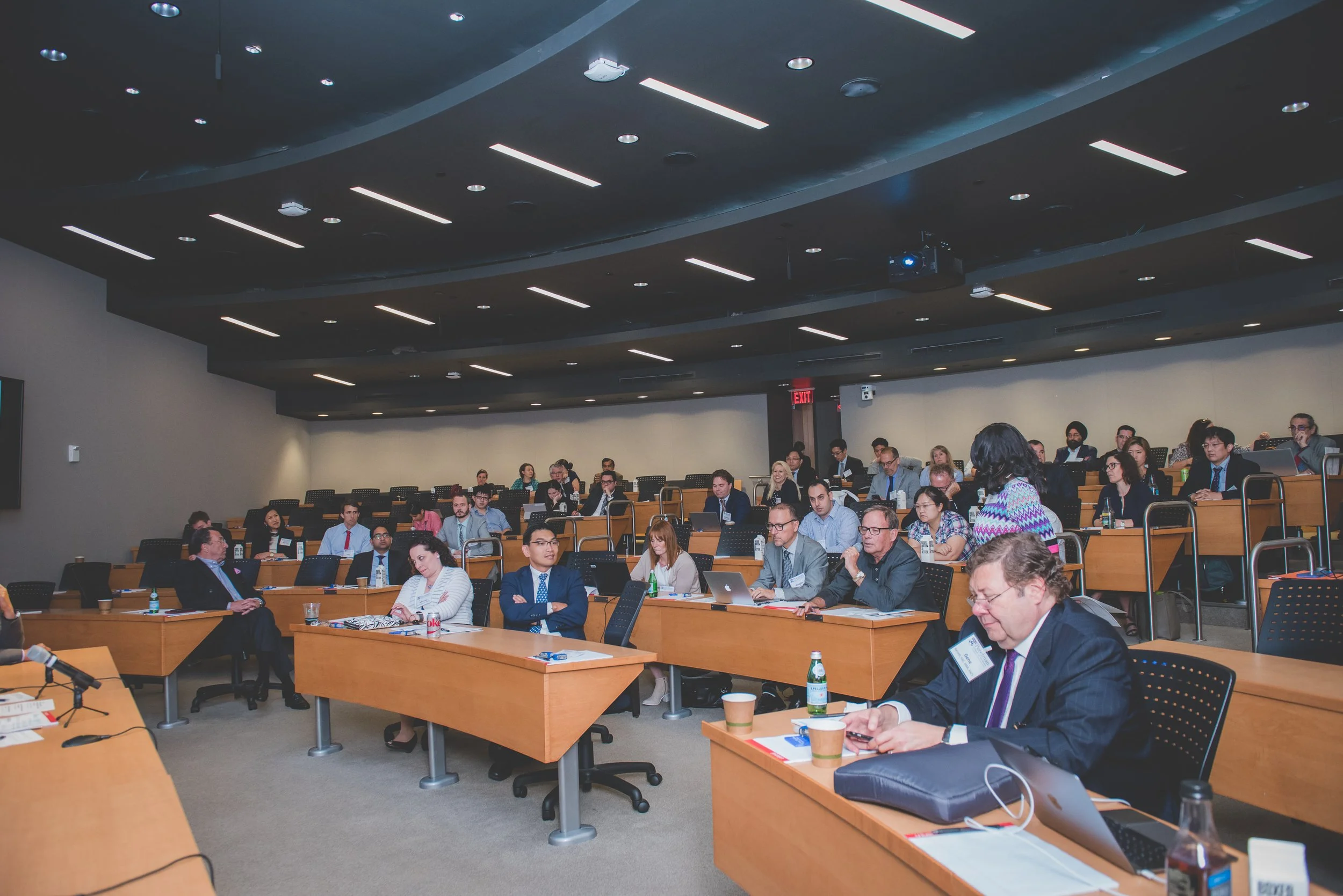 A large conference room filled with diverse attendees sitting at tiered tables, participating in a professional event or seminar.