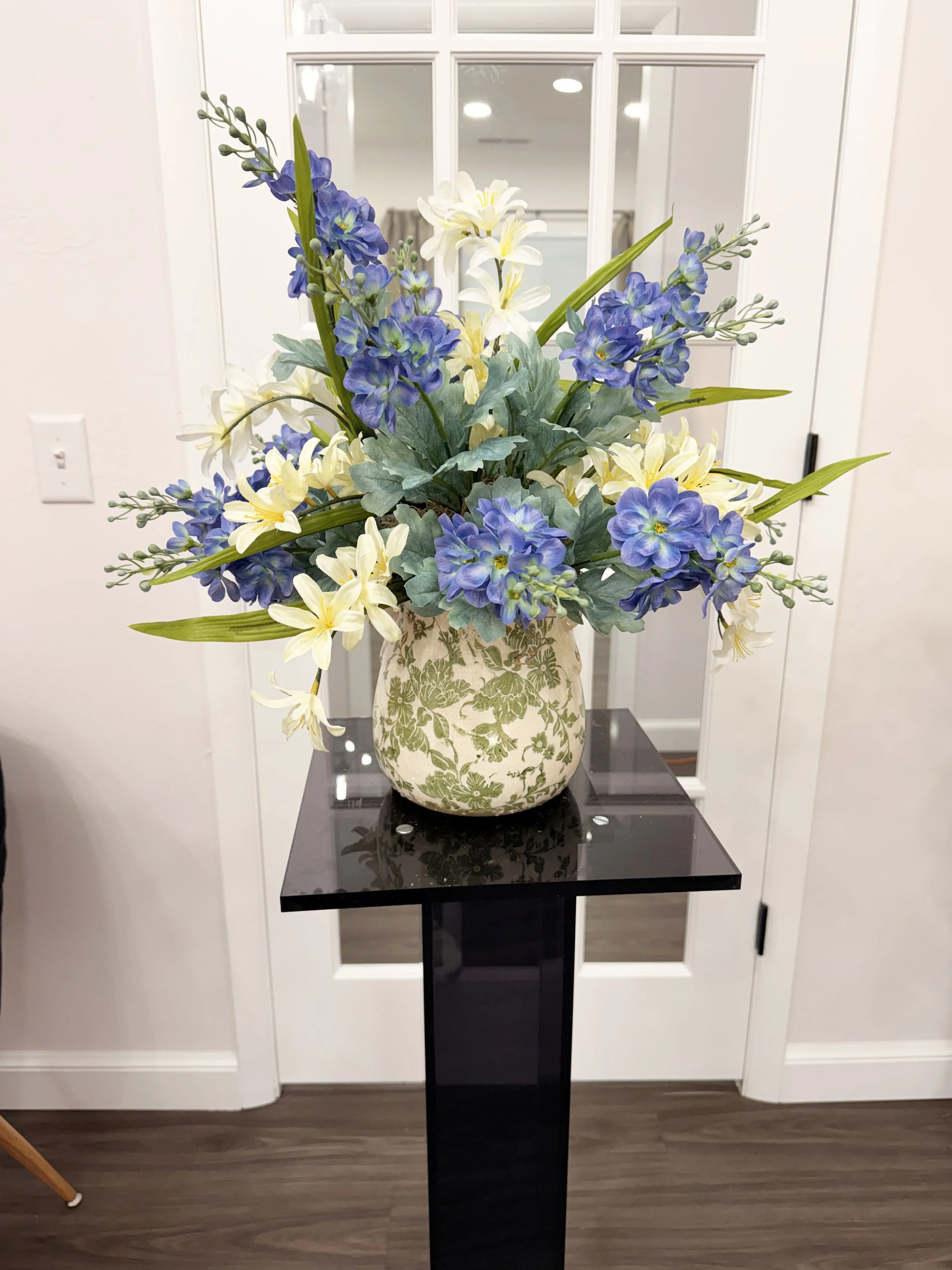 A floral arrangement with white, blue, and yellow flowers in a patterned ceramic vase placed on a black pedestal table in front of a white door with glass panes.