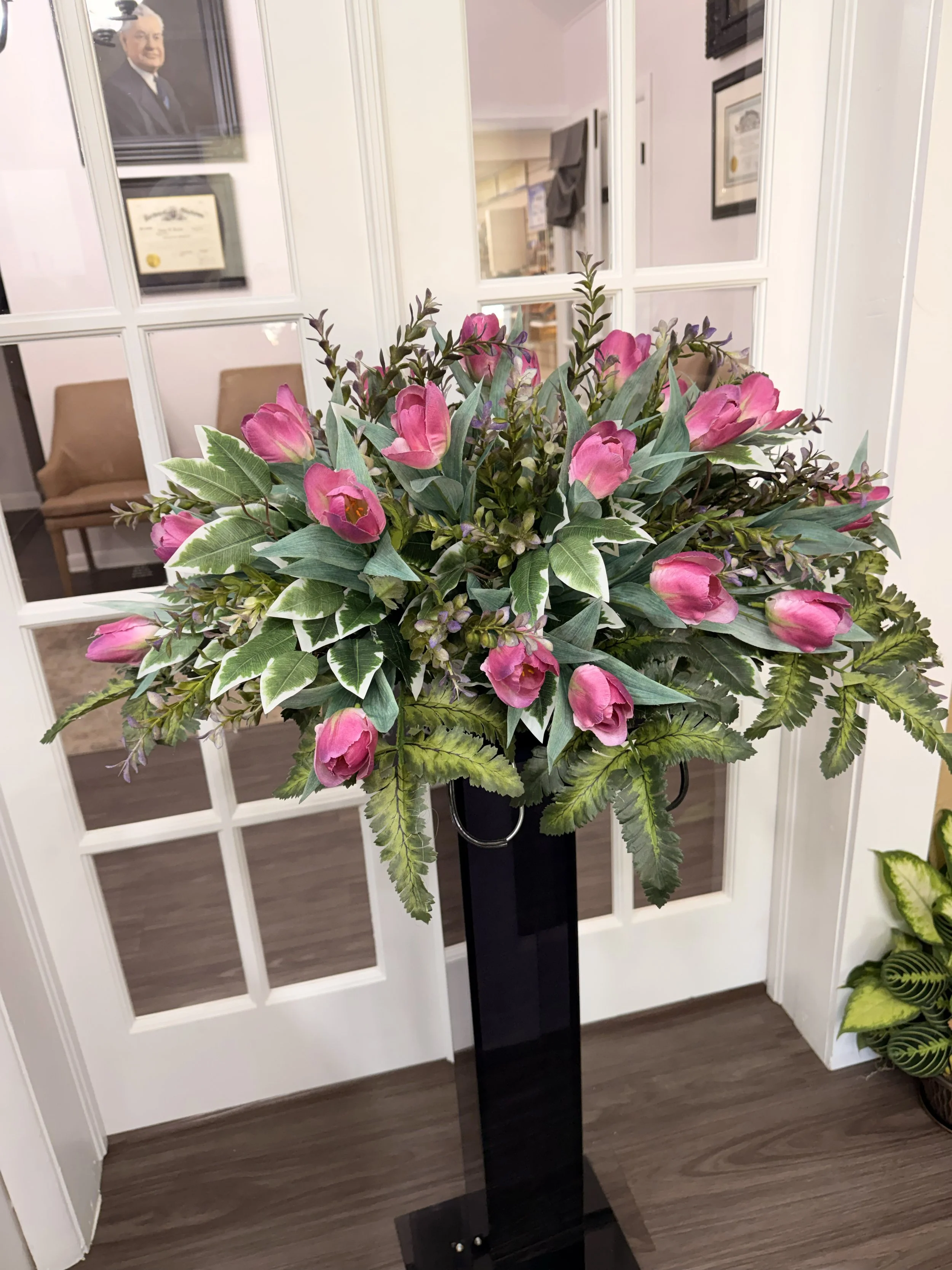 A large bouquet of pink flowers and green leaves is placed on a black stand in front of a glass-paneled door. Inside, a room with wall art and furniture is visible.