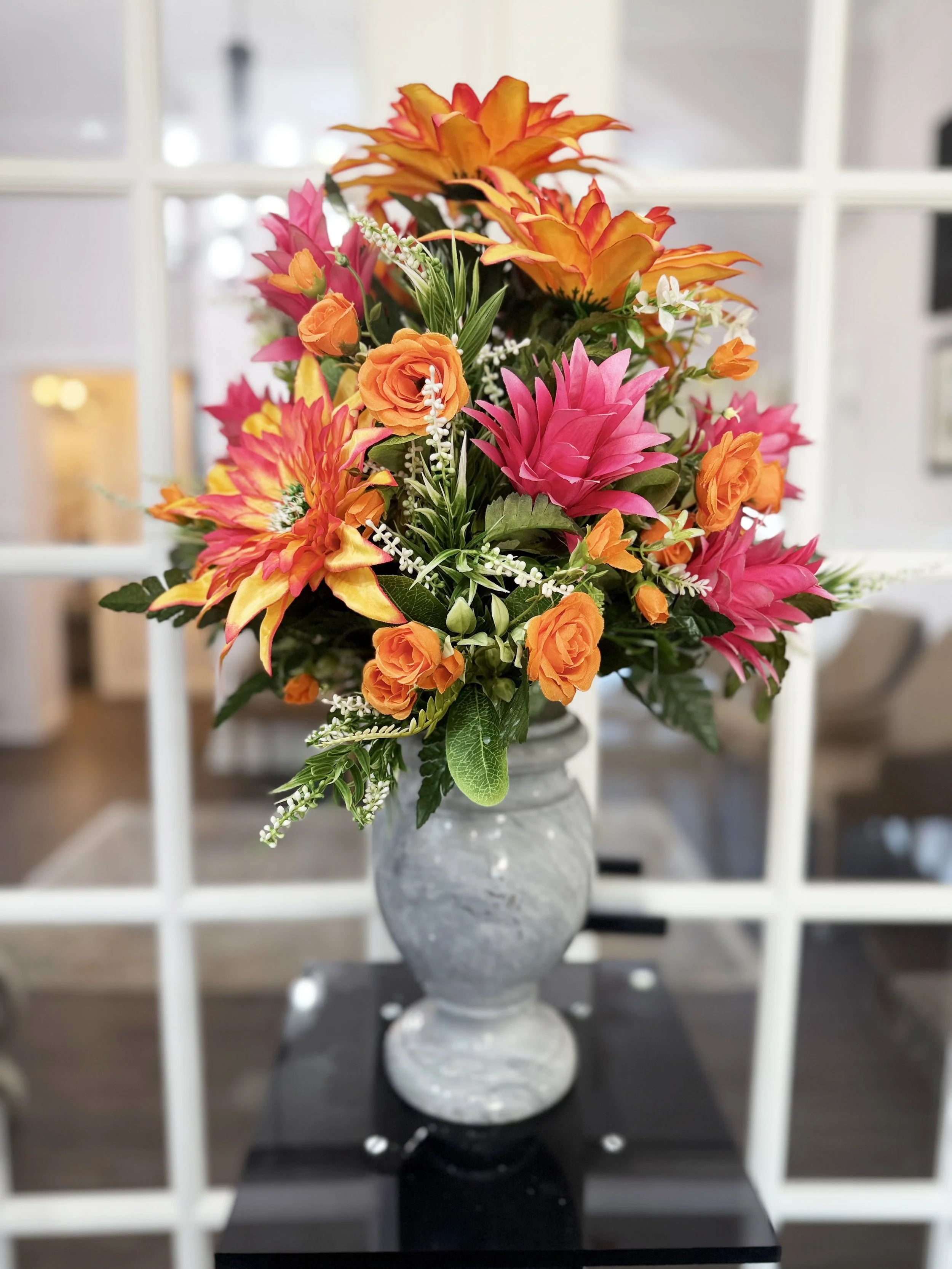 A colorful bouquet of flowers with orange roses, pink lilies, and white small flowers in a grey marble vase, placed on a black surface against a blurred background with white window panes.