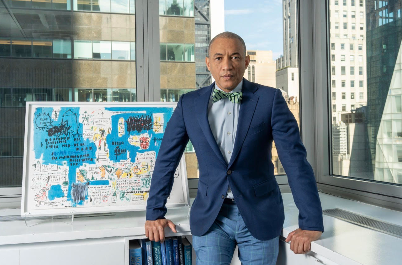 Man in blue suit with green bow tie standing in office with city skyline view, leaning on white desk with colorful drawings on whiteboard.