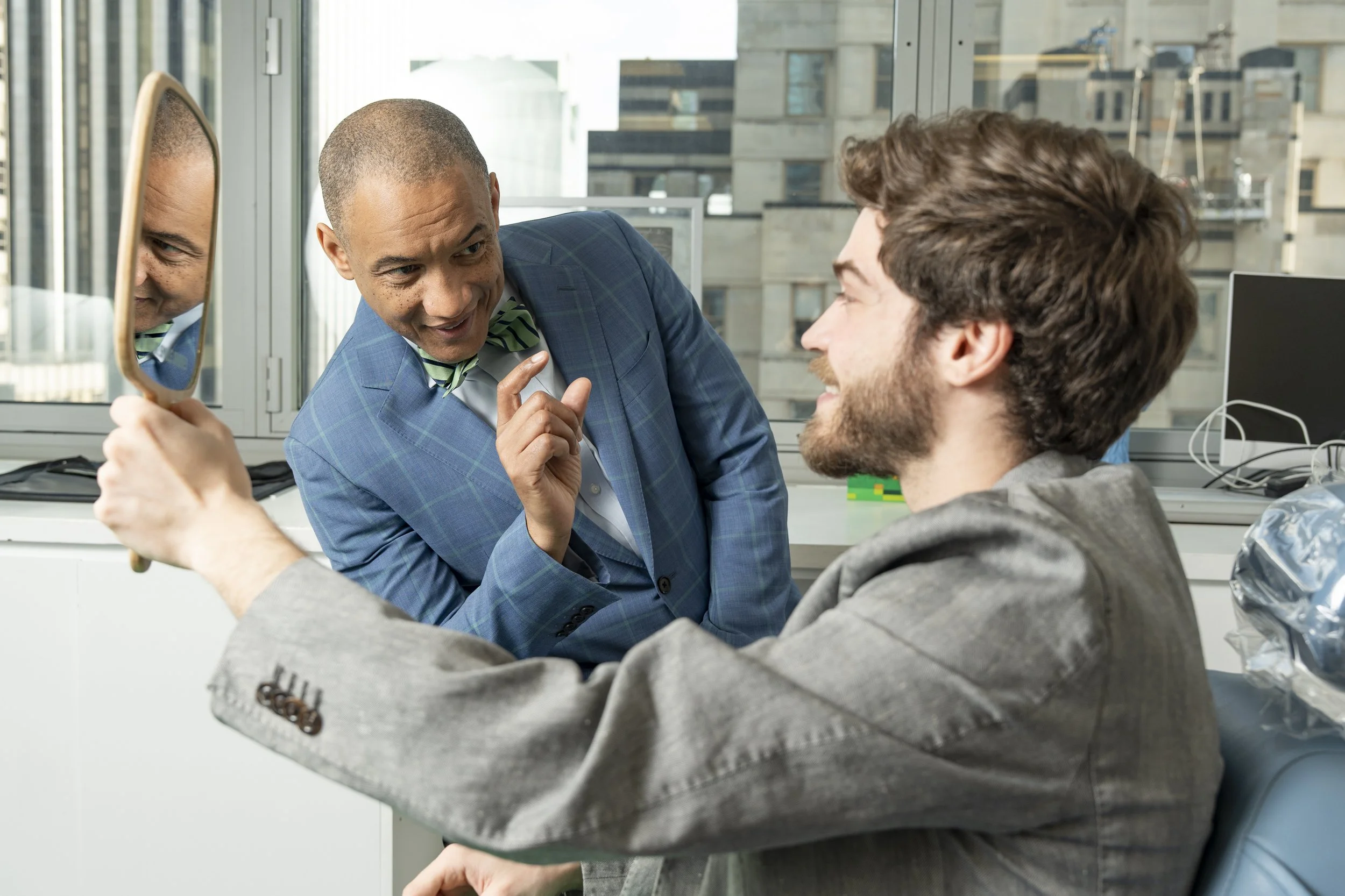 Two men in an office, one holding a mirror and smiling, the other pointing and talking, with city buildings outside the window.