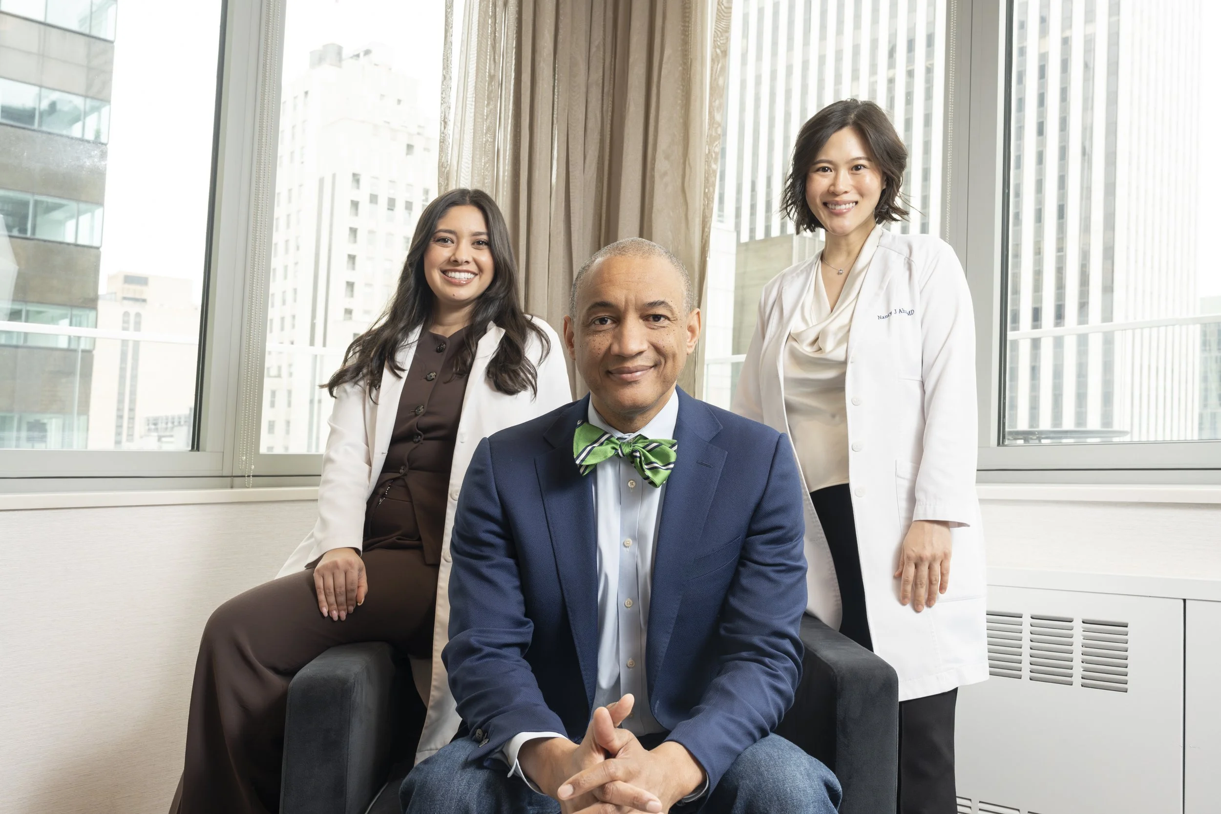 Three women and one man in an office with large windows and cityscape view, the man seated in front wearing a green bow tie, the women standing behind him smiling.