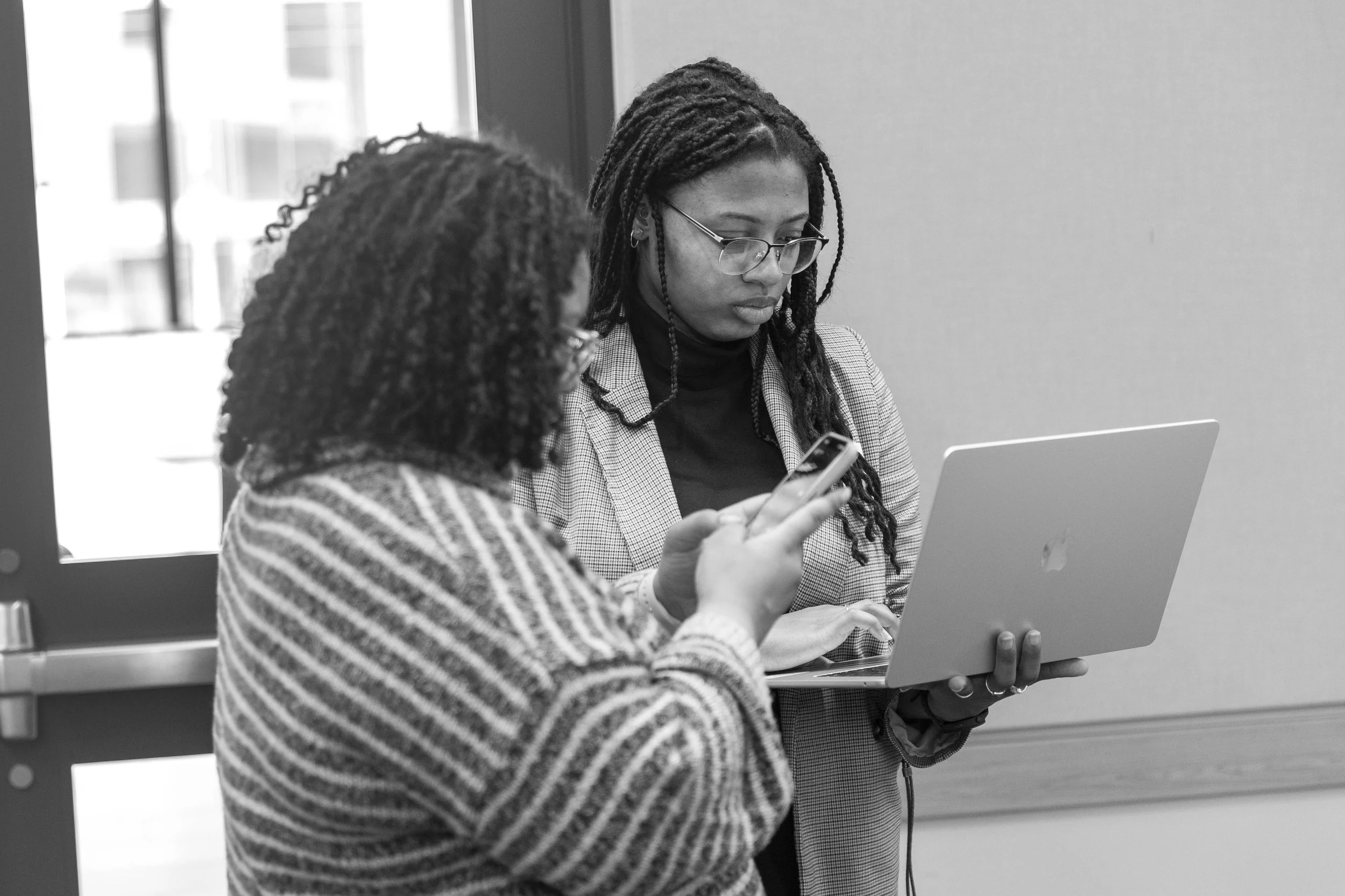 Two women with dreadlocks and curly dark hair, wearing glasses, sitting at a table with laptops and smartphones in an indoor setting.