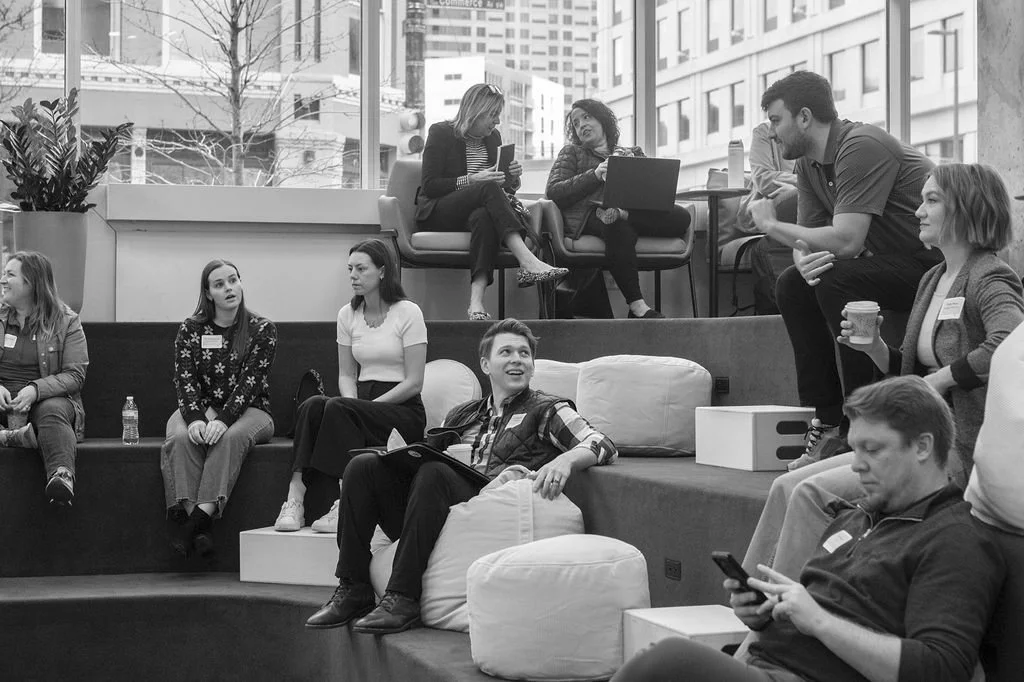 People sitting and standing in a modern office space, engaged in conversation, with some using laptops and phones, and a cityscape visible through large windows.